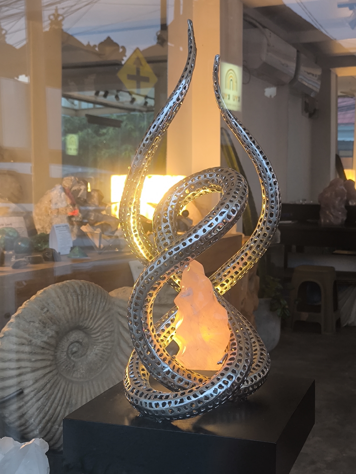 The media file displays a close-up view of a decorative lamp or sculpture, likely situated in a shop window, offering a layered composition of display items and reflections. The central subject is an intricate lamp featuring intertwining, perforated metallic elements that form an upward-spiraling design. Nestled within these silver-toned spirals is an illuminated, rough-hewn crystal, likely Himalayan salt, emitting a warm, amber-orange glow. This centerpiece rests on a black, rectangular display stand.
In the immediate background, to the left, a large, well-preserved ammonite fossil with prominent spiral patterns is visible on a lower shelf. Further into the shop, various other crystals and minerals are displayed on shelves, including green and grey specimens on the left and a larger pinkish crystal formation on the right.
The image is characterized by significant reflections on the glass, offering glimpses of the outdoor environment and parts of the interior not directly in the frame. Reflections show a yellow diamond-shaped road sign with a black cross and an upward arrow, suggesting a street intersection outside. An ornate architectural roofline, possibly indicative of traditional Indonesian design, is also reflected. High on an interior wall, air conditioning units are visible, along with a faint rainbow-colored arch logo. The lighting suggests it is daytime.
The setting appears to be a retail establishment specializing in minerals, fossils, and decorative items, consistent with the location context of North Kuta, Indonesia. No specific activity or people are depicted.