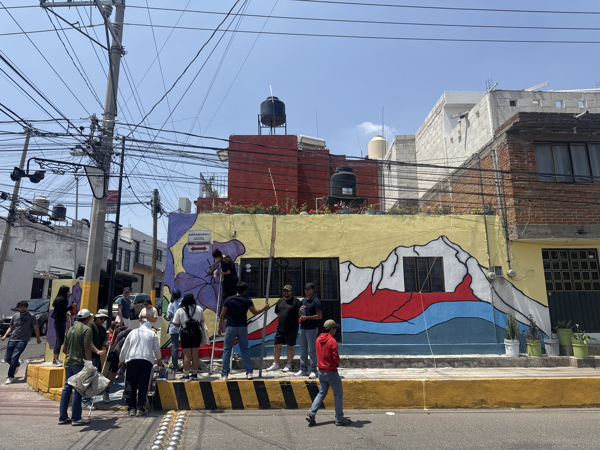 This vibrant image depicts a bustling street scene in Heroica Puebla de Zaragoza, Mexico, on a bright, sunny day. The central focus is a building adorned with a striking, colorful mural featuring stylized mountains in yellow, white, red, and blue.
A group of people, primarily adults, are gathered on the sidewalk in front of this building. Several individuals are actively engaged in painting or touching up the mural, with one person positioned on a ladder. Others are standing around, observing, conversing, or holding various items such as paint buckets, water bottles, and bags. The scene suggests a community art project or an organized painting event.
The urban setting is characterized by a dense network of overhead electrical wires strung between poles, a common sight in Mexican cities. The surrounding buildings showcase a mix of architectural styles, including unfinished concrete block structures and brick, with visible water tanks (tinacos) and solar water heaters on the rooftops.
Notable details include a yellow-painted curb with black diagonal stripes and embedded metal road reflectors. Visible text on signs includes "MATAMOROS" with directional arrows, along with smaller text like "SOMBRERIA" and "CANTINA." A partially visible commercial sign advertises "Coria," "PAPELERIA," "SCORES," and "24 HRS." The clear blue sky and bright sunlight indicate it is mid-day or early afternoon.