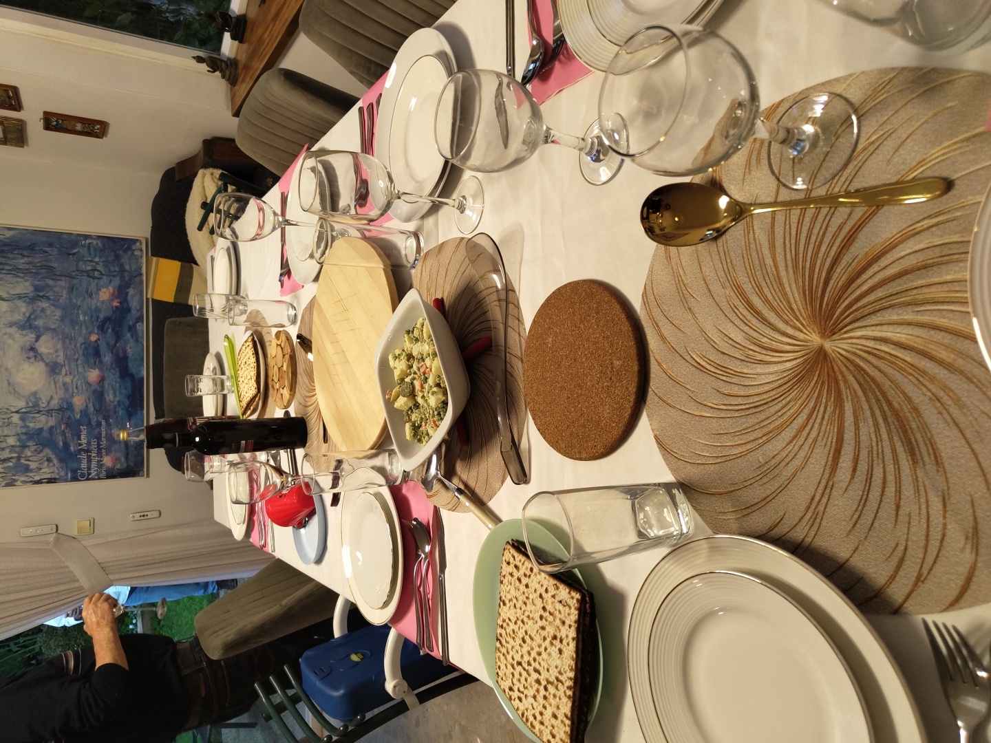 The image depicts a dining table set for a meal, likely a Passover Seder, in what appears to be a home in Raanana, Israel.
**Setting:**
* **Location:** Interior of a house, presumably in Raanana, Israel. The presence of Seder plate items suggests a Passover Seder.
* **Environment:** A table covered with a white tablecloth. Chairs are arranged around the table. A painting hangs on the wall and natural light enters from a window.
**People:**
* An adult can be seen in the lower left of the picture.
**Objects:**
* **Table setting:** Multiple place settings with plates, silverware, wine glasses, and water glasses.
* **Seder plate elements:** Matzah, Charoset, Karpas in a bowl.
* **Other:** Wine bottles, wooden cutting boards, coasters.
**Actions:**
* The table is set, indicating an impending meal.
**Interactions:**
* There are no active interactions visible. The scene is set and waiting for participants.
