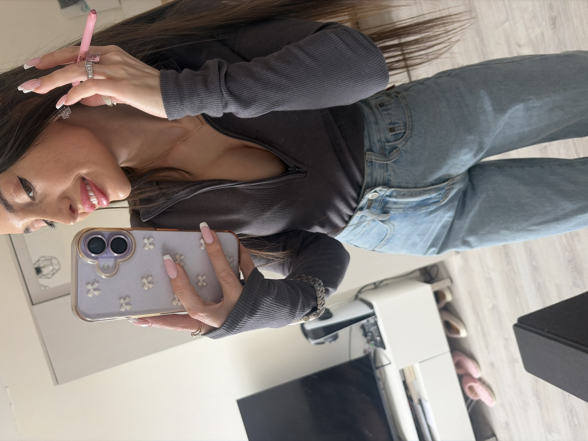 A young woman is taking a selfie indoors, angled from slightly above and to her left. She holds a lavender-colored phone case adorned with small white flowers in her left hand, which has manicured nails. She's wearing a charcoal gray zip-up top and blue jeans. A gold chain is visible around her neck. She also wears rings on her fingers, including one with a large pink stone. Her left hand also displays a silver watch or bracelet. Her long, dark hair falls over her shoulder.
In the background, there is a white cabinet or shelving unit with a dark TV mounted on the wall above it. Further behind the TV cabinet, the floor is visible. A couple of pairs of shoes are near the cabinet.
The image does not include any information to indicate the location as Saint Petersburg, Russia.
