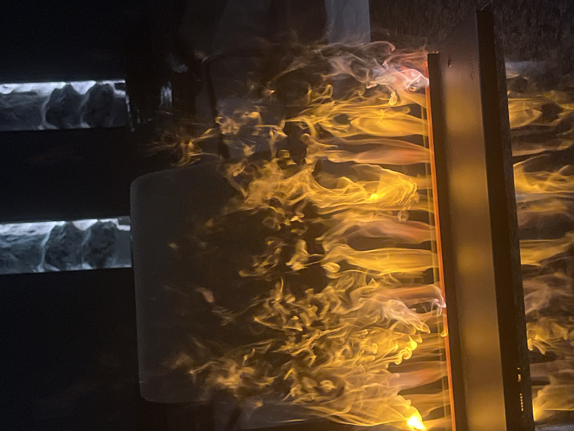 The image captures an indoor scene, likely a modern art installation or decorative architectural feature, set against a dark background. The central focus is a dynamic display of light and what appears to be simulated or real smoke/vapor, predominantly in vivid golden-yellow and orange hues.
On the right side of the frame, a vertical, dark panel frames a vibrant cascade of these glowing, fluid forms, resembling flames or billowing smoke. The light source appears to be behind or integrated into this structure, illuminating the moving patterns. The forms are intricately swirled and textured, creating a sense of constant motion.
To the left of this bright display, a more diffuse and ethereal area of similar golden-yellow and white smoke-like patterns is visible, suggesting either a continuation of the installation or a reflection on an adjacent surface. Further to the left and in the background, rectangular horizontal elements are visible. Within these dark frames, distinctive white and grey, wave-like or mountainous shapes are discernible, possibly another part of the same multi-faceted display or an abstract design. A lighter, possibly cylindrical or curved object is partially visible in the lower-left foreground.
The overall setting is dimly lit, emphasizing the glowing elements. The absence of natural light suggests an evening or nighttime interior. There are no identifiable people or visible text in the image. The scene, located in Sofia, Bulgaria, creates a mysterious and captivating ambiance through its interplay of light, shadow, and fluid forms.