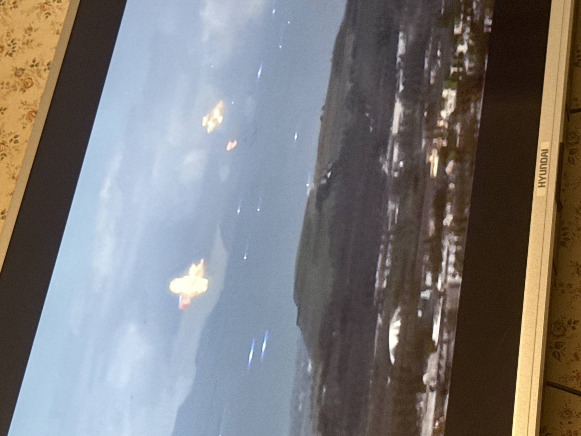 A photo of a Hyundai monitor displaying what appears to be an aerial view of Mytishchi, Russia, possibly captured from a video game. Two distinct explosions with bright, fiery centers are visible against a backdrop of sky and landscape. Streaks of light suggest the presence of projectiles or falling debris. A hill or mountain range flanks one side, while the urban landscape of Mytishchi stretches along the other. The resolution is low, resulting in a blurred image.