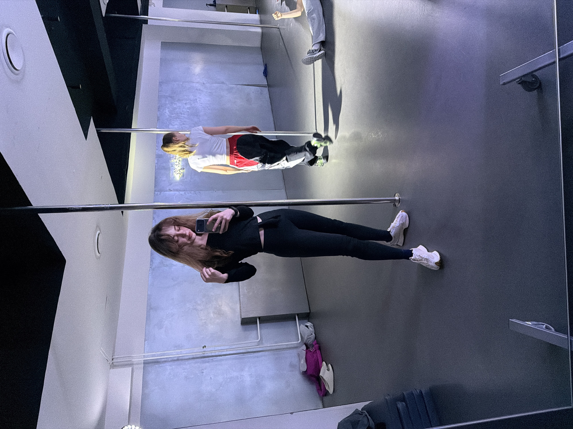 This image captures an indoor pole fitness studio located in Moscow, Russia. A young female with long brown hair, dressed in a black long-sleeved crop top and black leggings, is visible in the foreground, reflected in a large mirror that forms the left wall. She is holding a dark-colored smartphone to take a selfie, with her right hand near her hair. She wears white athletic shoes. Behind her, also reflected, another young female with blonde hair tied in a ponytail wears a white crop top and black, red, and white track pants, standing near a chrome vertical pole. A third individual is partially visible lying on the dark grey floor in the far right background, wearing grey sweatpants and sneakers, with one arm raised. The studio features concrete-textured walls, one displaying a yellow neon sign with Cyrillic text, and exposed white piping. The dark grey floor reflects ambient light and the studio's features. On the left side of the floor, a stack of dark mats, a pair of light-colored slip-on shoes, and a pile of pink and grey fabric items are visible. The ceiling is white with recessed lighting and black structural elements.