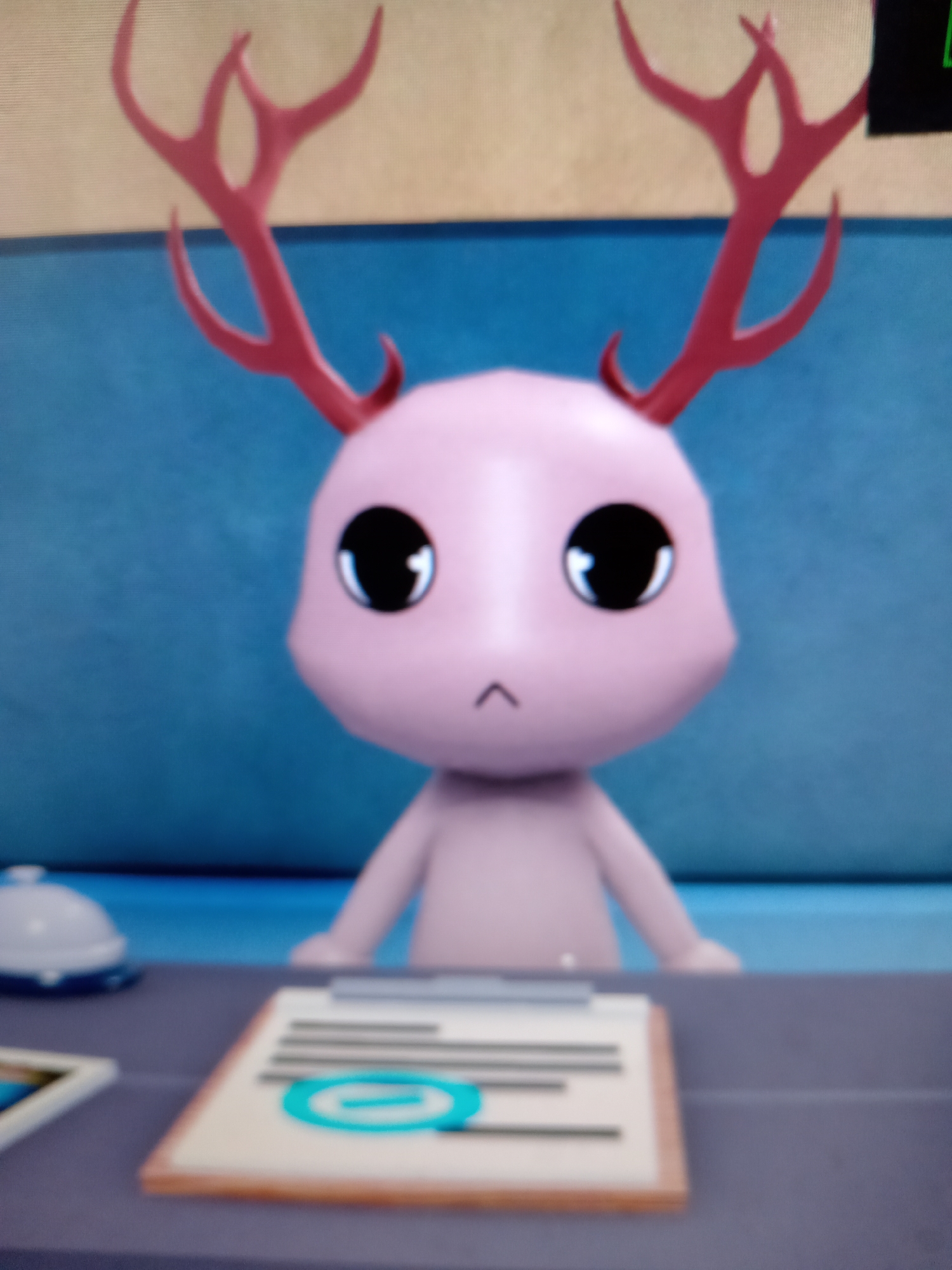 A pink, cartoonish creature with large black eyes, red antlers, and no discernible mouth is centered in the frame, looking forward. It appears to be sitting behind a grey desk. On the desk, to the left, is a white bell. To the right of the bell is a clipboard with papers and a blue circular symbol. The background is a textured blue wall with a beige strip across the top. The image is of low resolution and appears to be a screenshot from a video game or animation. The location of Rudny, Kazakhstan is not discernible from the image.