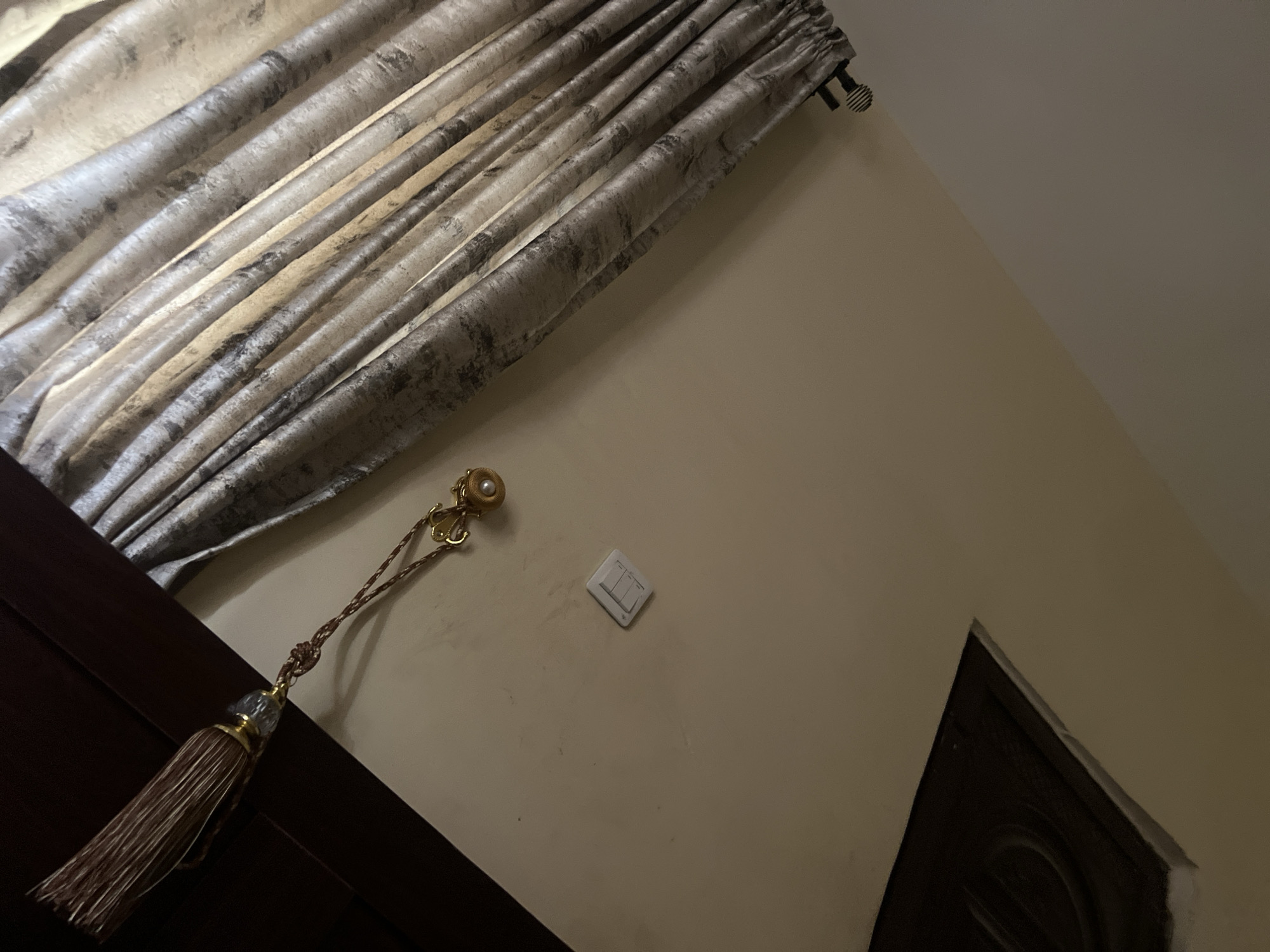 This image presents an indoor scene, likely a room in a residence in Howayi, Nigeria. The upper portion of the frame is dominated by pleated curtains, made of a light-colored fabric, possibly beige or off-white, with a mottled or abstract greyish-brown pattern. These curtains hang from a dark curtain rod, visible on the top right.
Below the curtains, a light-colored wall, appearing cream or light beige, spans most of the visible area. Mounted diagonally on this wall is a white, rectangular light switch with two toggle buttons. To the left of the light switch, a decorative golden-colored curtain tie-back or tassel is prominently featured. It includes a pearl-like accent, a braided cord, and a multi-stranded light brown tassel.
In the bottom left corner, a dark brown, possibly wooden, structure is partially visible, suggesting a piece of furniture such as a bed frame or a wardrobe. Similarly, a dark brown door or window frame with some carved detailing is seen partially in the bottom right corner.
The lighting in the image appears dim, consistent with an interior setting, possibly during evening hours or in a room with limited natural light. There are no people visible, and no specific activities are taking place. No visible text is present in the image.