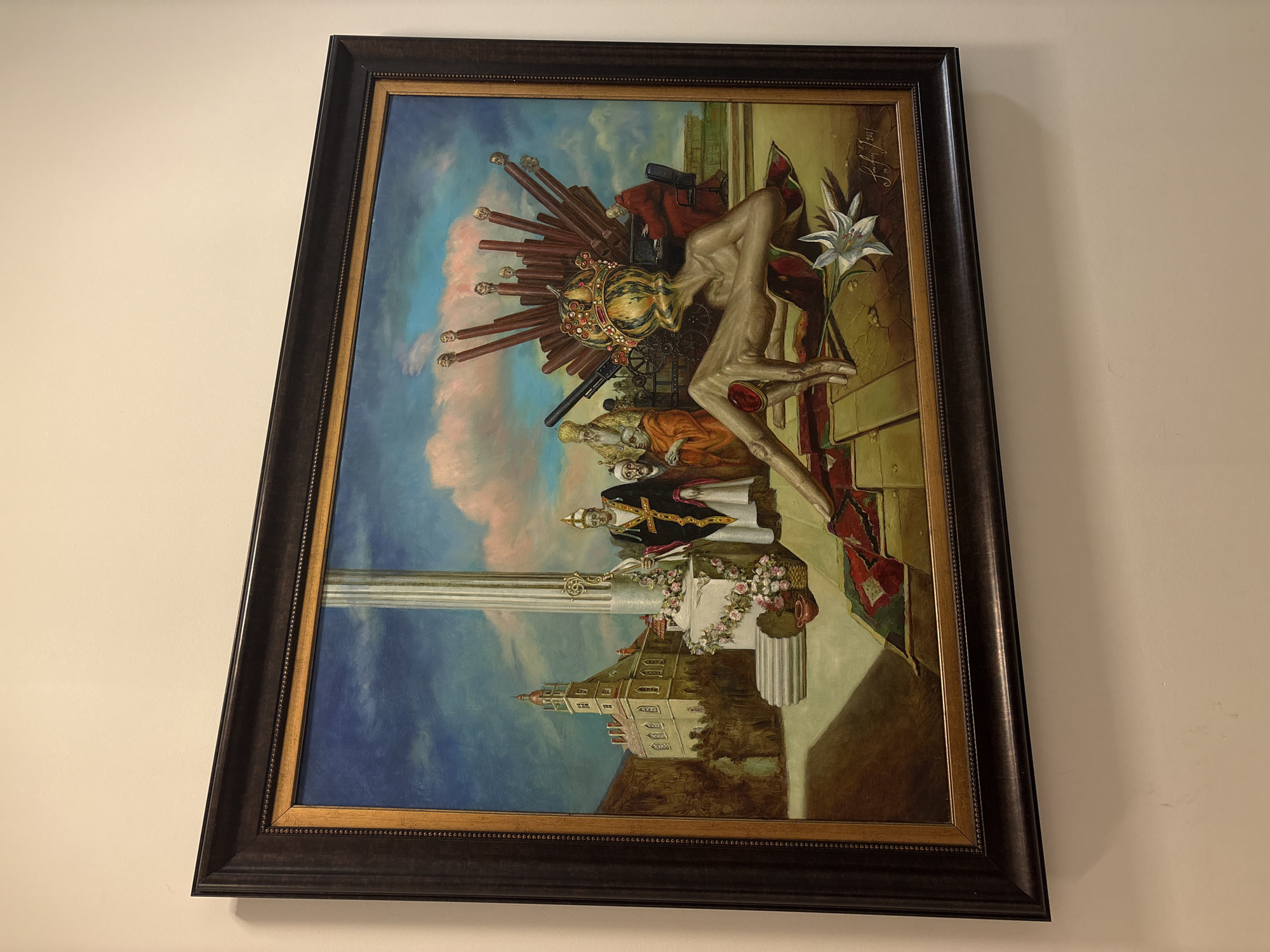The image shows a framed painting hanging on a light beige wall. The painting depicts a surreal scene. The lower part of the painting includes a building resembling a church or palace, topped with what appear to be flowers and possibly figures in clerical attire. Above them is a column and a complex scene with multiple figures. At the top is an abstract figure wearing a crown with multiple protruding objects, and a lily or similar flower beside it. There is a gun or other weapon depicted in the middle of the composition. I am unable to determine an exact location within Moscow based on the painting.