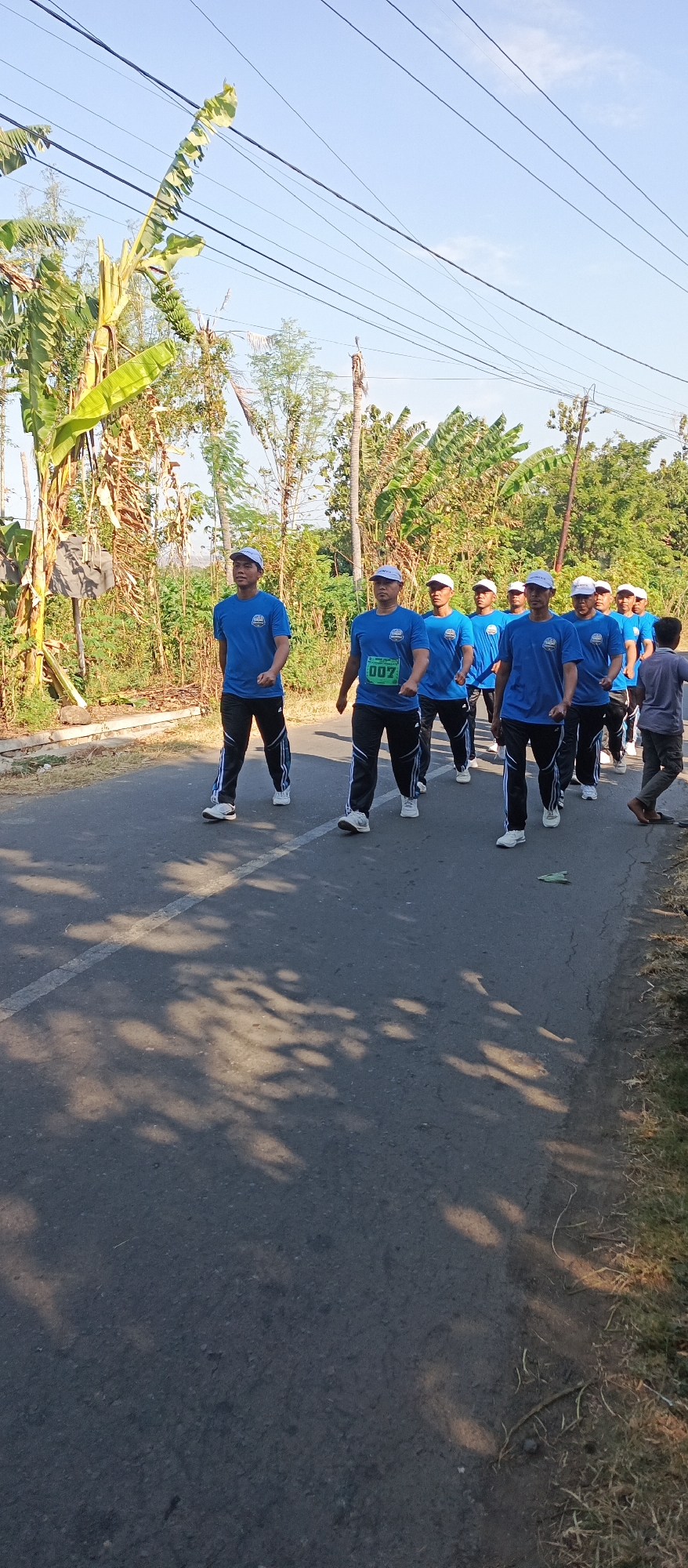 In Ketowan, Indonesia, a group of men in matching blue t-shirts and white caps are walking in single file on a paved road. The men appear to be participating in some sort of organized event, possibly a walk or march, as indicated by their uniform attire and the presence of a bib with the number "007" on the shirt of one of the men. The road is bordered by lush vegetation, including banana trees, and power lines are visible overhead. The sun is shining, casting shadows on the road, suggesting it is daytime. The overall impression is one of an outdoor community event or activity taking place in a semi-rural or suburban setting.