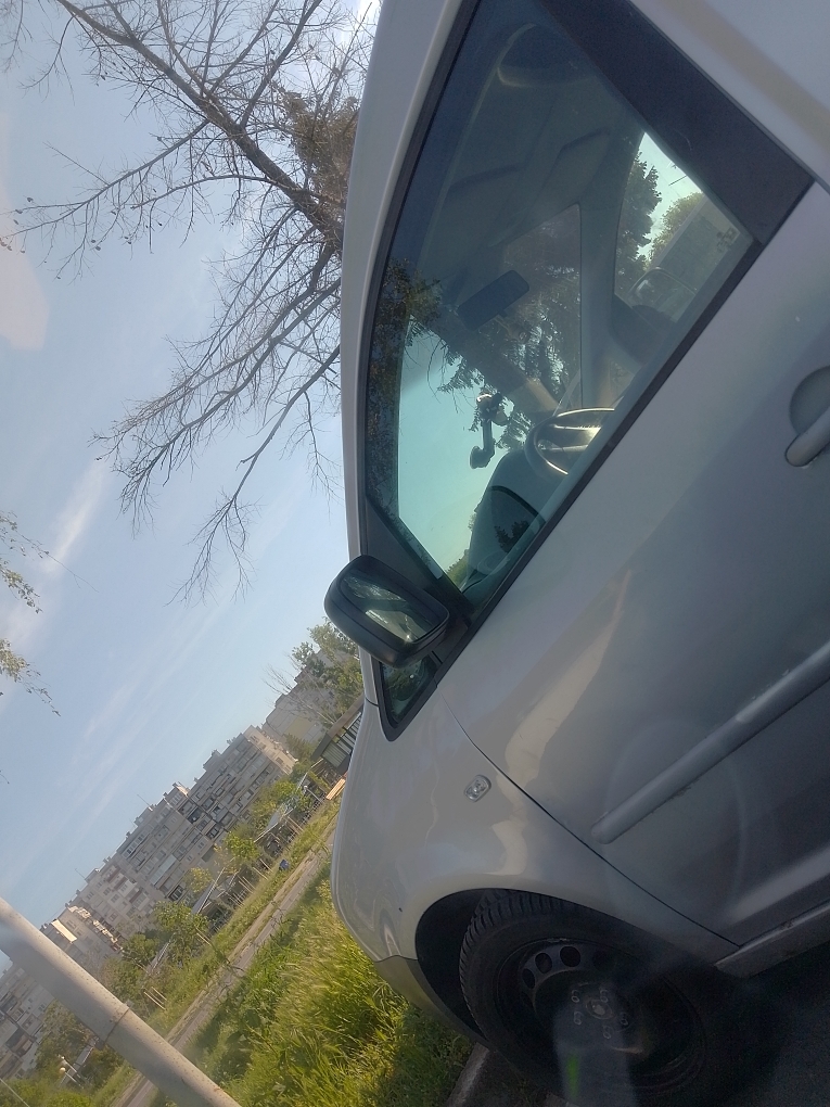 A silver car is parked on the side of a street, angled towards the right. The car's front tire is visible, along with the side door and mirror. Reflected in the car window are the car's interior and a tree's branches. In the background, there is a row of apartment buildings and a grassy area with trees. The sky is light blue. The location appears to be in a city.
