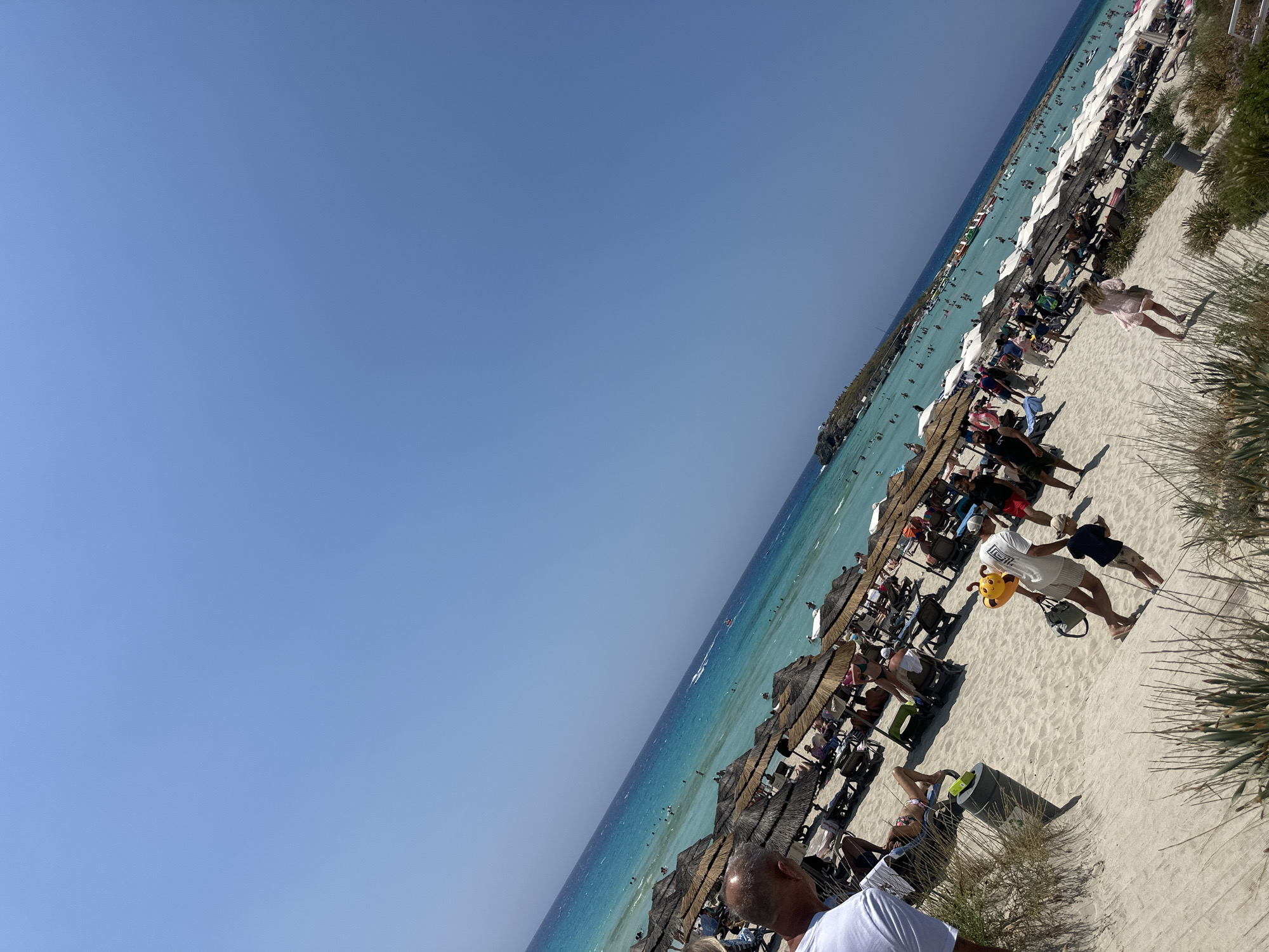 The image depicts a bustling beach scene in Ayia Napa, Cyprus. The sand is light-colored and stretches towards the turquoise waters of the Mediterranean Sea. Numerous beach umbrellas with thatched roofs and white canopies are set up, providing shade for beach chairs and loungers occupied by people. The sea is populated with swimmers, and several watercraft, including what appears to be a jet ski, are visible. In the foreground, people are walking along the sandy shore, some carrying bags and one man is holding a yellow toy. The sky is a clear, vibrant blue, indicating a sunny day. The overall impression is one of a popular tourist destination during peak season.