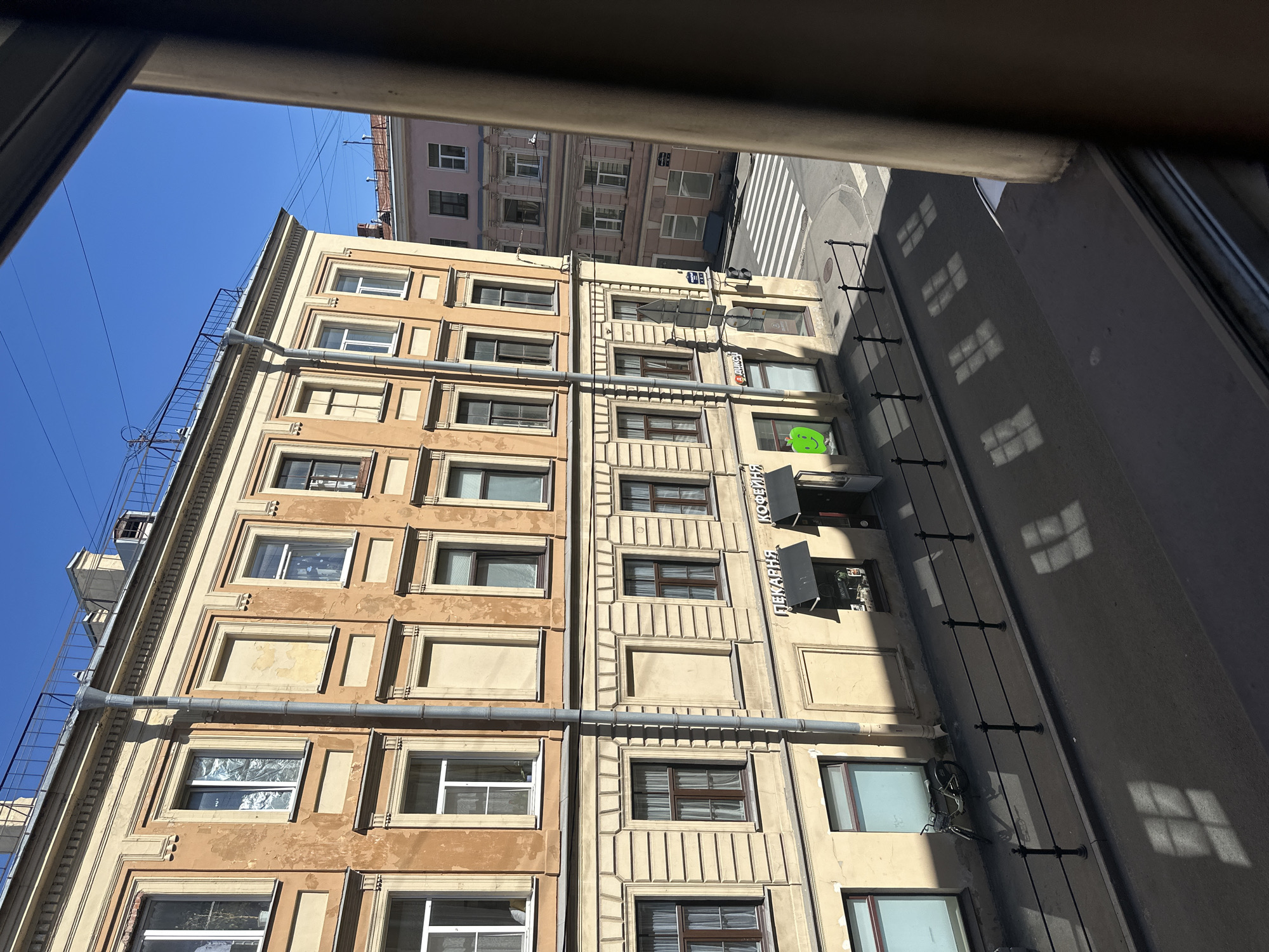 The image depicts a street scene in the city of Unknown, Unknown, viewed from an elevated window on a clear, sunny day. The bright blue sky is visible at the top. The immediate foreground is framed on the right by a dark window frame and along the bottom by a dark window sill.
Across the street, a multi-story building occupies the left and central portions of the frame. Its facade presents a two-tone appearance: a cream-colored lower section with horizontal textured patterns and an upper section painted a faded, peeling orange-brown. The building features multiple rectangular windows; some have white frames and appear open, others have darker frames and are closed, and several are visibly boarded up or stuccoed over. A grey metallic drainpipe runs vertically along the building's front. The rooftop is equipped with a dark metal railing and various antennas or structures.
At the ground level of this building, commercial establishments are present. One shop entrance is covered by a black awning and displays signs in white Cyrillic script reading "ПЕКАРНЯ" (Bakery) and "КОФЕЙНЯ" (Coffee Shop). Another storefront features a large, bright green apple logo on its window. A partially obscured sign in Cyrillic for "ДИКСИ" is also visible. A dark-colored bicycle is parked upright against the building on the sidewalk.
The asphalt street in the middle ground curves sharply to the right, leading to an intersection marked by a white zebra pedestrian crossing. A series of black metal bollards or railings delineates the sidewalk from the street in the foreground. Strong, rectangular shadows, cast by unseen windowpanes, are prominently visible on the street pavement, indicating a low sun angle.
At the intersection on the right, a second multi-story building with a light pinkish-beige facade and multiple windows is visible. Overhead, several utility or communication cables stretch across the clear sky. No people or moving vehicles are present in the scene.