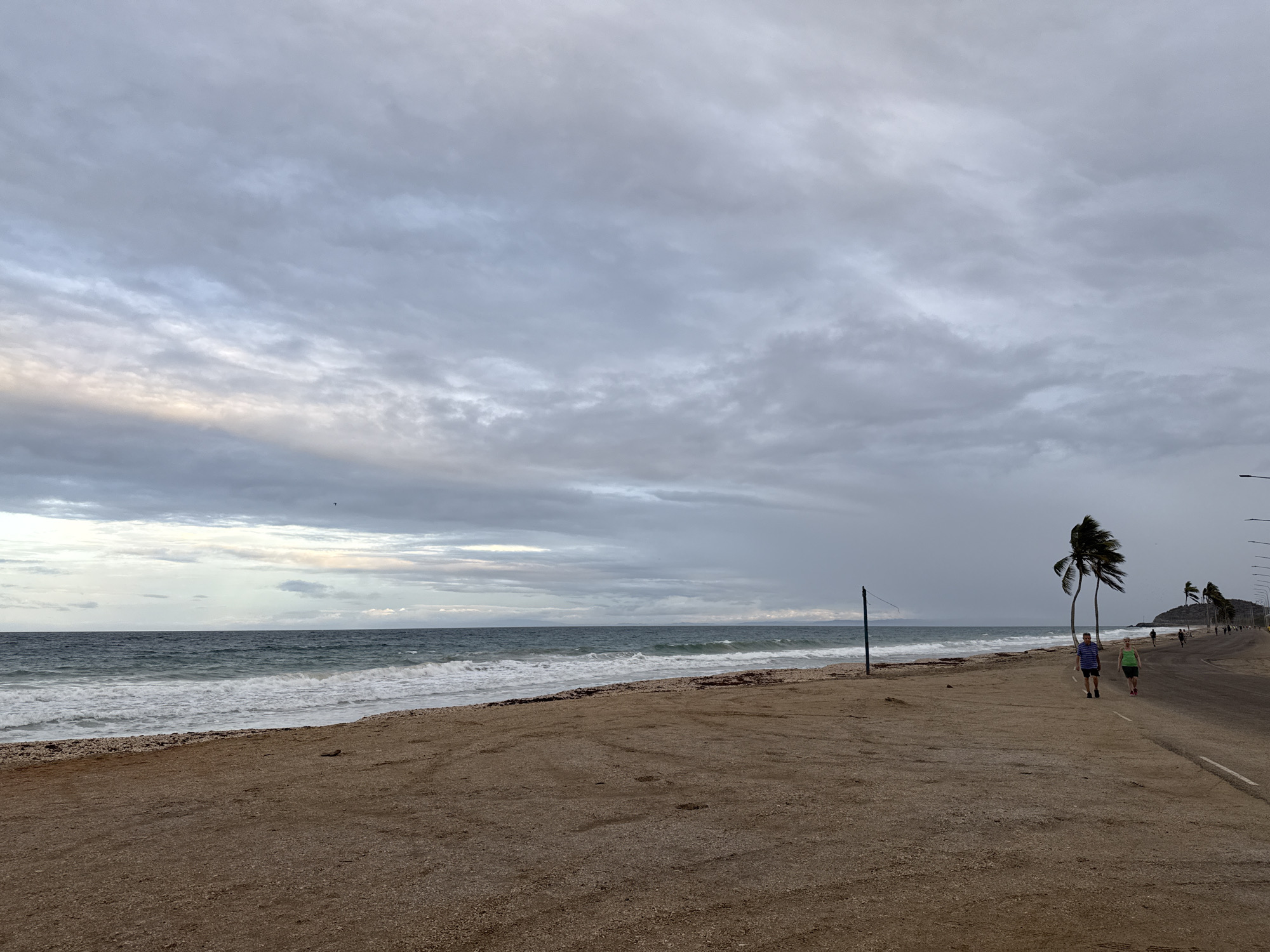 This wide-angle photograph captures an overcast coastal scene in Porlamar, Venezuela, encompassing a beach, the ocean, and a paved road running parallel to the shore. The sky is largely covered by thick, grey clouds, with a faint strip of lighter, diffused light visible near the horizon on the left, hinting at either early morning or late afternoon.
The foreground consists of a broad stretch of sandy and pebbly beach, showing some tire tracks. The ocean, appearing in dark blue-green hues, features white-capped waves breaking onto the shore. In the mid-ground, a tall, slender blue pole, possibly a post for a volleyball net or a marker, stands on the sand closer to the waterline.
Two individuals are walking on the sandy shoulder adjacent to the paved road. The person on the left wears a striped blue and white shirt with dark shorts, while the person on the right is dressed in a bright green top and dark shorts, both appearing to be walking casually.
To the right, a paved road with a white lane marker extends into the distance, lined by several tall palm trees with fronds appearing to sway in the wind. Further along the road, more people can be seen in the distance, their forms silhouetted. Utility poles or streetlights also flank the road. Faint landmasses are visible across the water in the far background. The overall mood is quiet and subdued due to the cloudy weather. There is no visible text in the image.