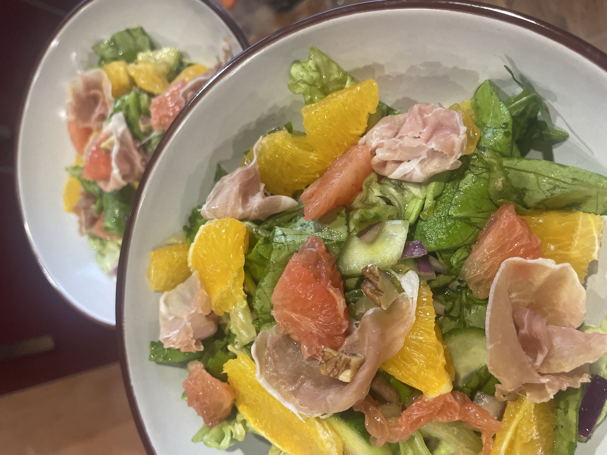 Two bowls of salad are presented. Each bowl contains a mix of lettuce, orange segments, pink grapefruit segments, and folded slices of pale pink meat (likely prosciutto). Other ingredients may be present, but are less visually prominent. The bowls have a light-colored interior and a brown-rimmed edge. The background is slightly blurred, suggesting a shallow depth of field. There is no indication of the location being Burgas, Bulgaria, or any other specific place.