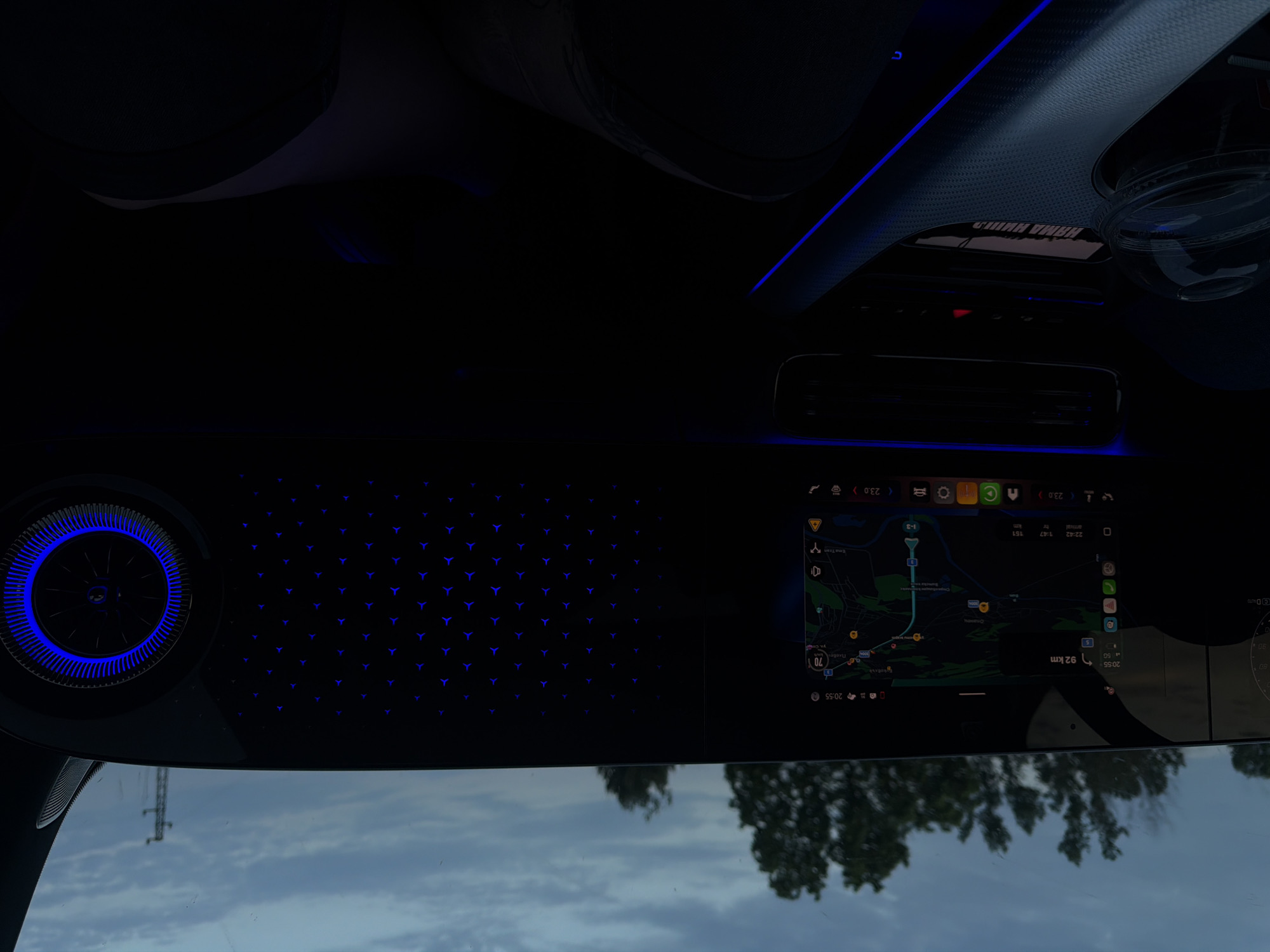 The image captures the interior of a vehicle at twilight, with a view through the windshield. The prominent feature is the vehicle's dashboard, which houses a large digital display. The left portion of this display shows a partial speedometer or gauge cluster. The central section functions as a navigation system, displaying a map with roads and geographical features. Text visible on the screen includes "20:55," "92 km," "5G" network indicator, a "70 km/h" speed limit icon, and estimated arrival information: "22:42" (arrival time), "1:47" (duration), and "151 km" (distance). The map clearly shows the Cyrillic text "Крушовица" (Krushovitsa) and "Плевен" (Peleben), indicating a location within or en route to Krushovitsa, Bulgaria. Below the map, icons indicate a temperature setting of "23.0" degrees Celsius.
To the right of the navigation screen, the dashboard features a textured panel embedded with a grid pattern of illuminated blue Mercedes-Benz star logos, serving as ambient lighting. Further to the right, a circular air vent is visible, framed by an illuminated blue ring.
Below the main display, the center console is visible. It features blue ambient light strips running along its contours. A textured, dark surface covers parts of the console. A clear plastic cup is present in a cup holder on the left side of the console. No occupants are discernible within the vehicle.
Through the windshield, the sky outside is overcast and dark, indicative of evening or dusk. Silhouettes of trees are visible against the sky, and a utility pole or transmission tower stands on the far right horizon.