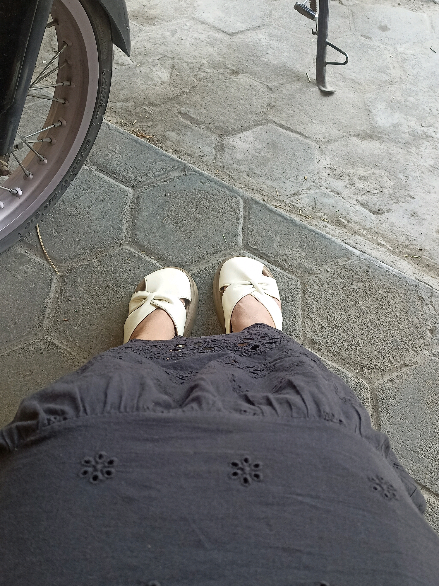 A first-person perspective shot of a person wearing a dark grey skirt with embroidered floral cutouts and white slip-on sandals with a bow detail. The person is standing on a paved area with hexagonal grey stones. To the left of the frame is the wheel of a motorcycle. A motorcycle kickstand is visible in the upper right corner of the image. The scene is likely outdoors in a public or residential area within Gabungan, Indonesia, given the paving style and presence of a motorcycle.