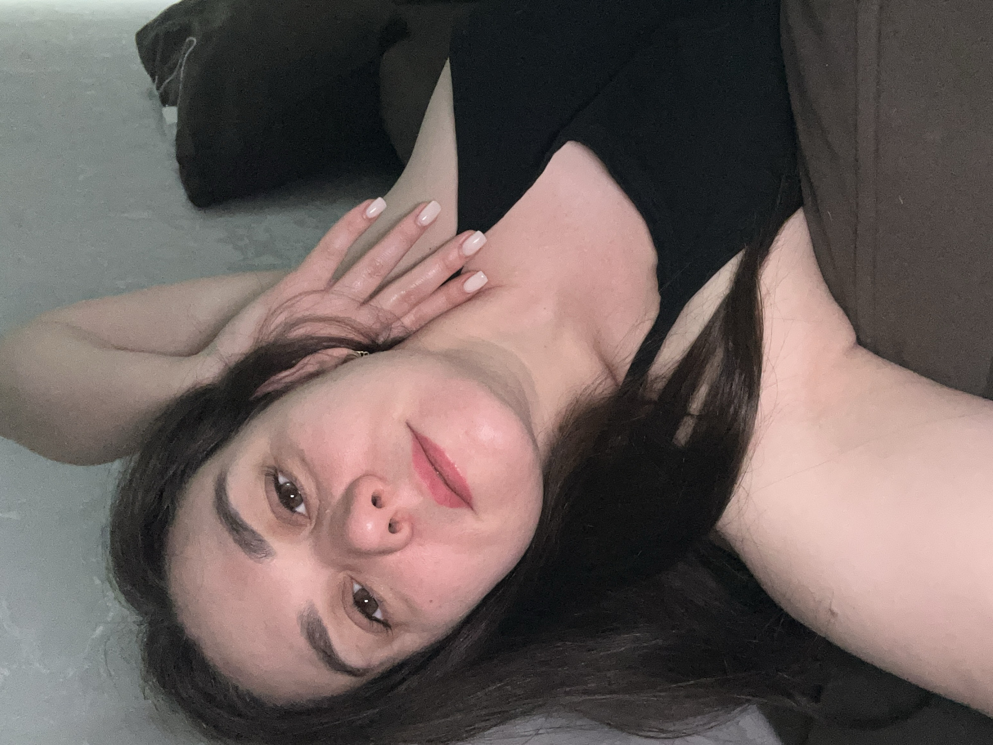 A woman with dark hair is lying down. She is wearing a black tank top. She is indoors on a grey surface with a dark brown pillow or couch behind her. Her right hand is placed on her chest/neck area. The photo does not provide enough context to determine her location in Chaikovsky, Russia.