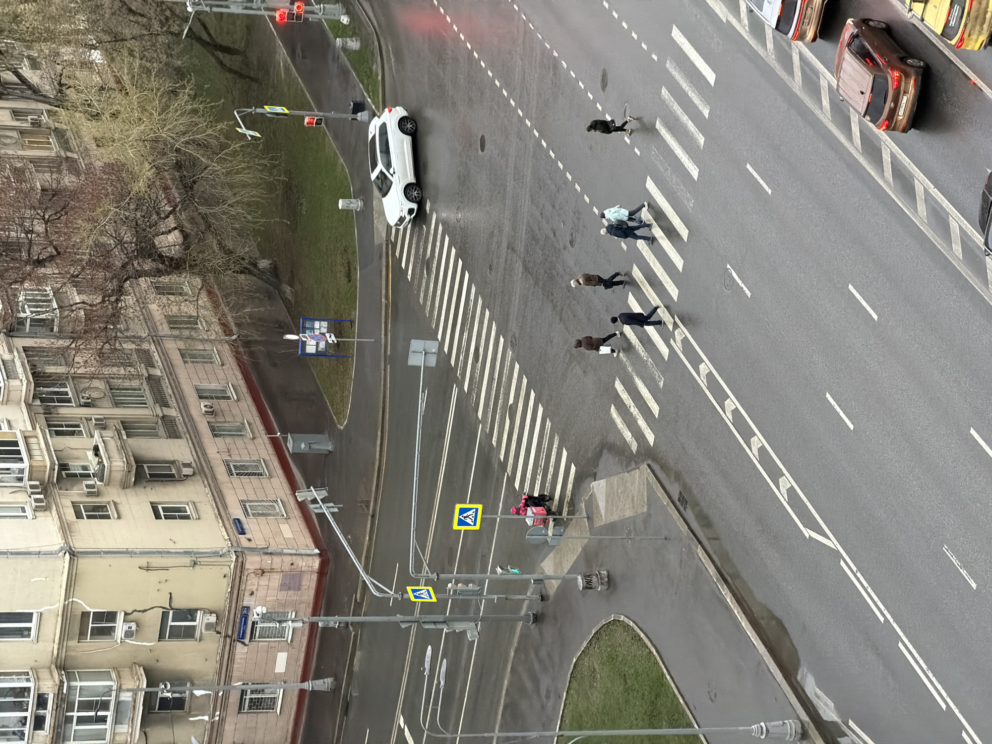 The image depicts a street scene in Moscow, Russia, from an elevated perspective. The scene includes pedestrians crossing the street at a zebra crosswalk. There are cars parked along the right side of the street, and a white car is turning. Streetlights and pedestrian crossing signs are visible on the left side of the street. Buildings are visible in the background. The sky is overcast, and the street appears wet.