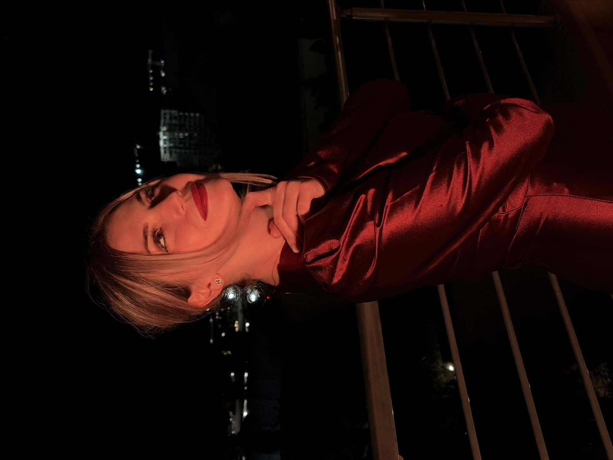 A woman is posing for a photo at night on a balcony in Budva, Montenegro. She is wearing a red, satin dress and has her right hand on her chin, looking up and to the right. The background shows a dimly lit cityscape with some buildings and lights visible. The lighting on the woman is warm, suggesting artificial light.