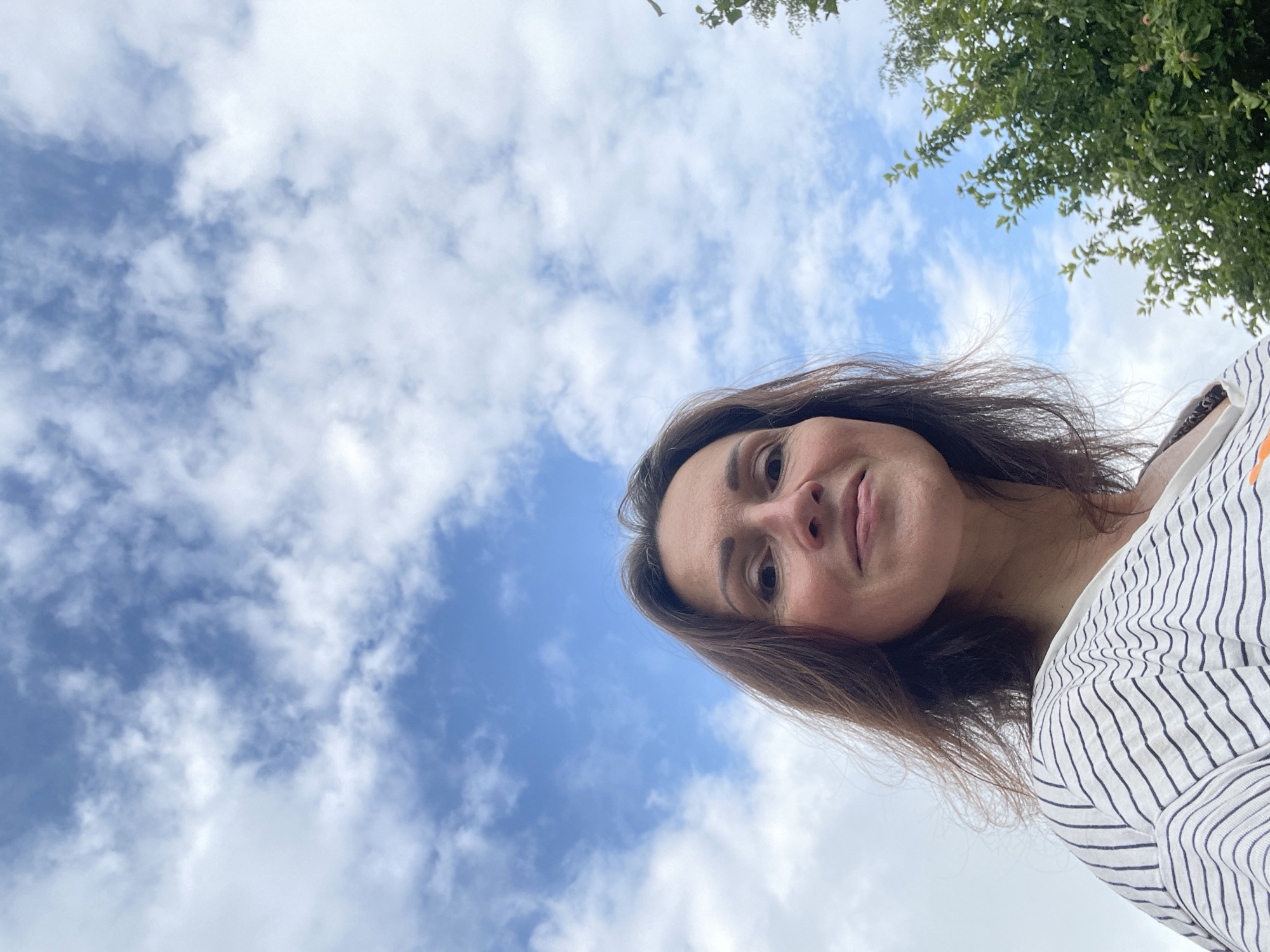 An outdoor, low-angle, medium shot features a woman from the chest up, looking towards the camera. She has long, dark brown hair, dark eyes, and is smiling. She is wearing a white t-shirt with thin, horizontal black stripes. The background is a bright blue sky with scattered white clouds. On the right side of the image, a green tree with dense foliage is visible. The image appears to have been taken in Дресвяново, Russia, on a cloudy day.