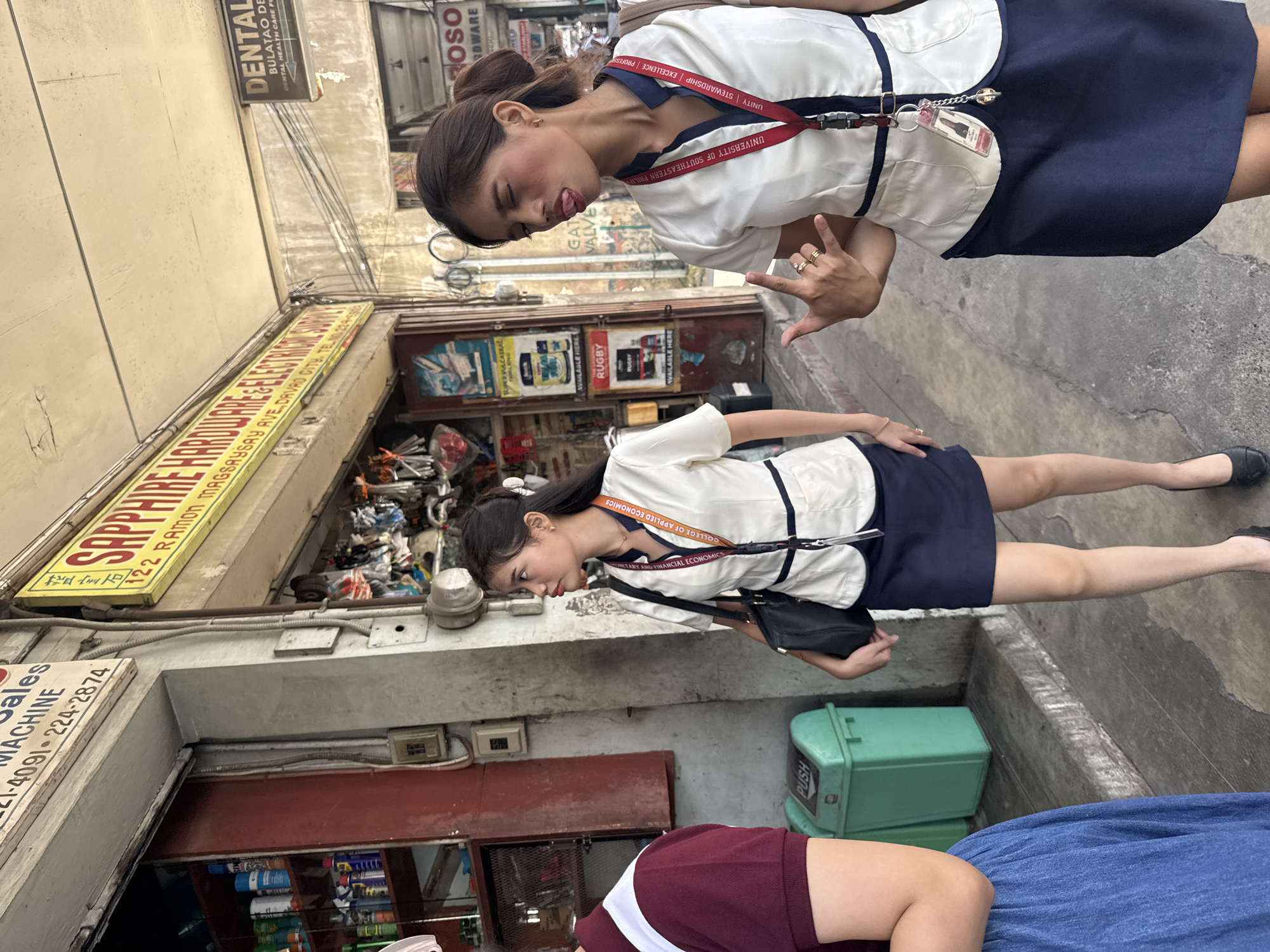 In an outdoor urban setting, likely a commercial street in Davao City, Philippines, two young women in uniform walk on a sidewalk. The woman on the right, positioned closer to the camera, playfully sticks out her tongue and makes a rock-on hand gesture. Both women wear white collared blouses with navy blue trim and matching navy blue skirts. They are also adorned with lanyards bearing the insignia "COLLEGE OF APPLIED ECONOMICS" and "UNIVERSITY OF SOUTHEASTERN PHILIPPINES". Behind them, the facades of several businesses are visible, including "SAPPHIRE HARDWARE & ELECTRICAL SUPPLY" with an address and phone number, and a sign for "DENTAL". Shelves stocked with various products are seen through open shop doorways. A green trash bin with a "PUSH" label is present on the left side of the image. The ground is paved with concrete and wooden planks. The overall atmosphere suggests a casual outing or a break from their academic activities.