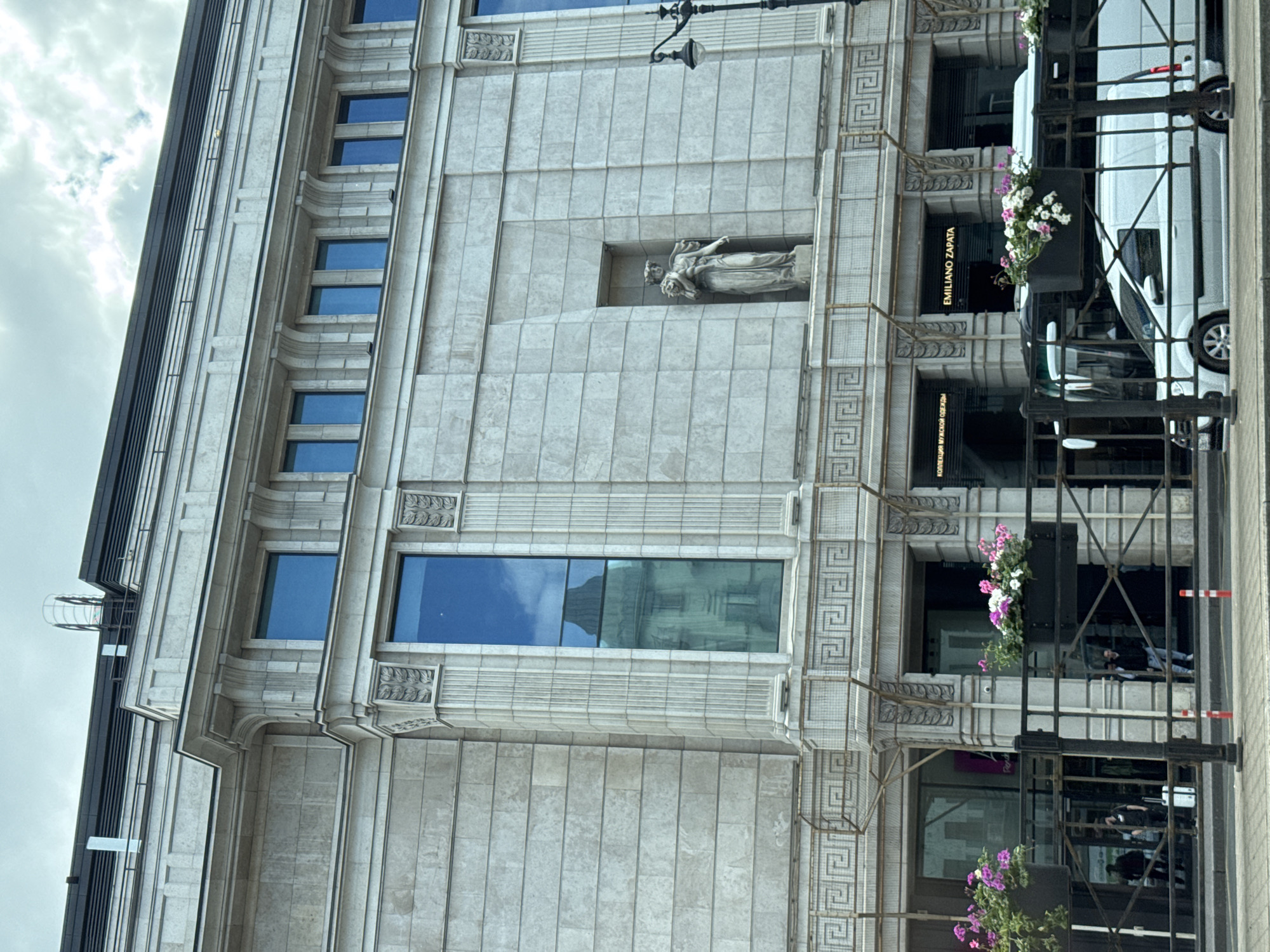 The image shows a multi-story building with a neoclassical facade in Saint Petersburg, Russia. The building is made of light-colored stone blocks and features decorative elements such as columns, cornices, and recessed niches. A statue of a female figure holding a bouquet is visible in one of the niches. Large, tall windows are present, some reflecting the sky and other buildings. In front of the building, there is a sidewalk with a black metal fence. Planters with pink and white flowers are mounted on the fence. Several people are seen walking in the background, and there are two vehicles, a white van and a bus, on the street in front of the building. Signs with text, including "EMILIANO ZAPATA," are visible above some of the building's entrances. The sky is overcast.