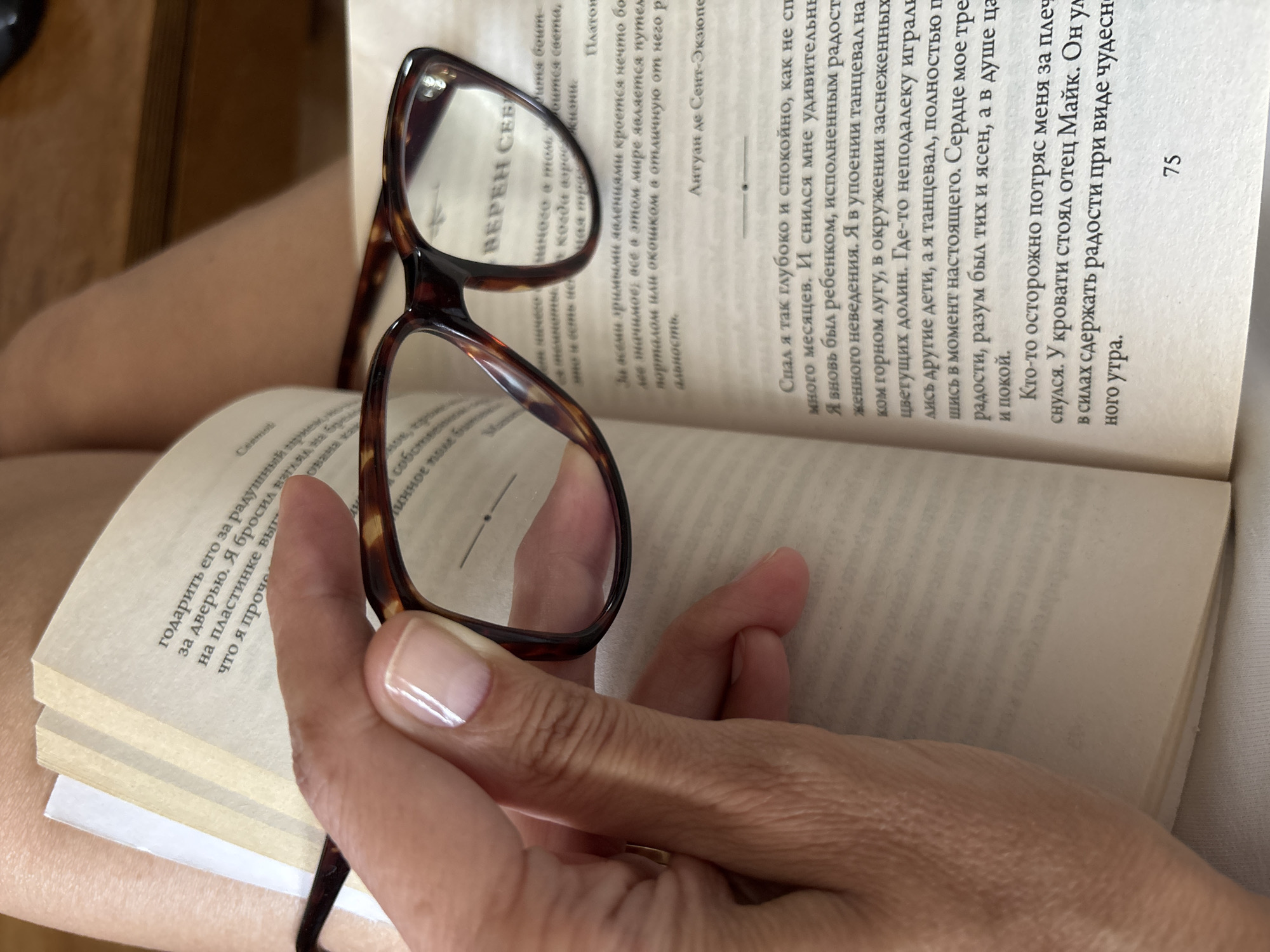 A photograph shows a book open on a person's lap, with a pair of eyeglasses held in front of the text. The person's hand, with manicured fingernails, is visible holding the glasses. The book's pages are filled with text in a Cyrillic alphabet, and the number "75" is visible on the right-hand page. The text discusses themes of sleep, childhood, and inner peace. The location is not discernible from the image.
