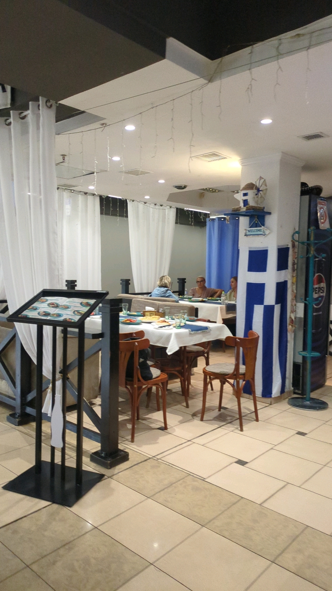The image shows the interior of a Greek-themed restaurant, likely located in Chișinău, Moldova. White and blue drapes and a large Greek flag decorating a pillar suggest a Mediterranean theme. Several tables are set for dining, with white tablecloths, plates, and glasses. In the foreground, a black stand displays a menu. In the background, three individuals are seated at tables, engaged in conversation. A Pepsi vending machine is visible on the right side of the frame. The floor is tiled with a light-colored pattern.