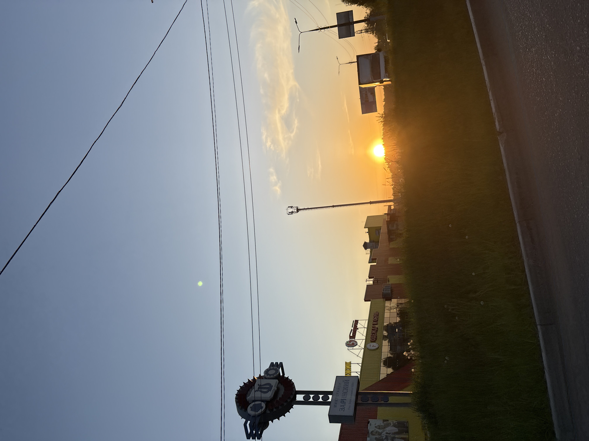 The image depicts a sunset over the city of Chaikovsky, Russia. A road and grassy area are visible on the right, with several buildings in the background. Power lines stretch across the sky from left to right. In the foreground is a sign with Cyrillic text, indicating "Zarinsky microdistrict."