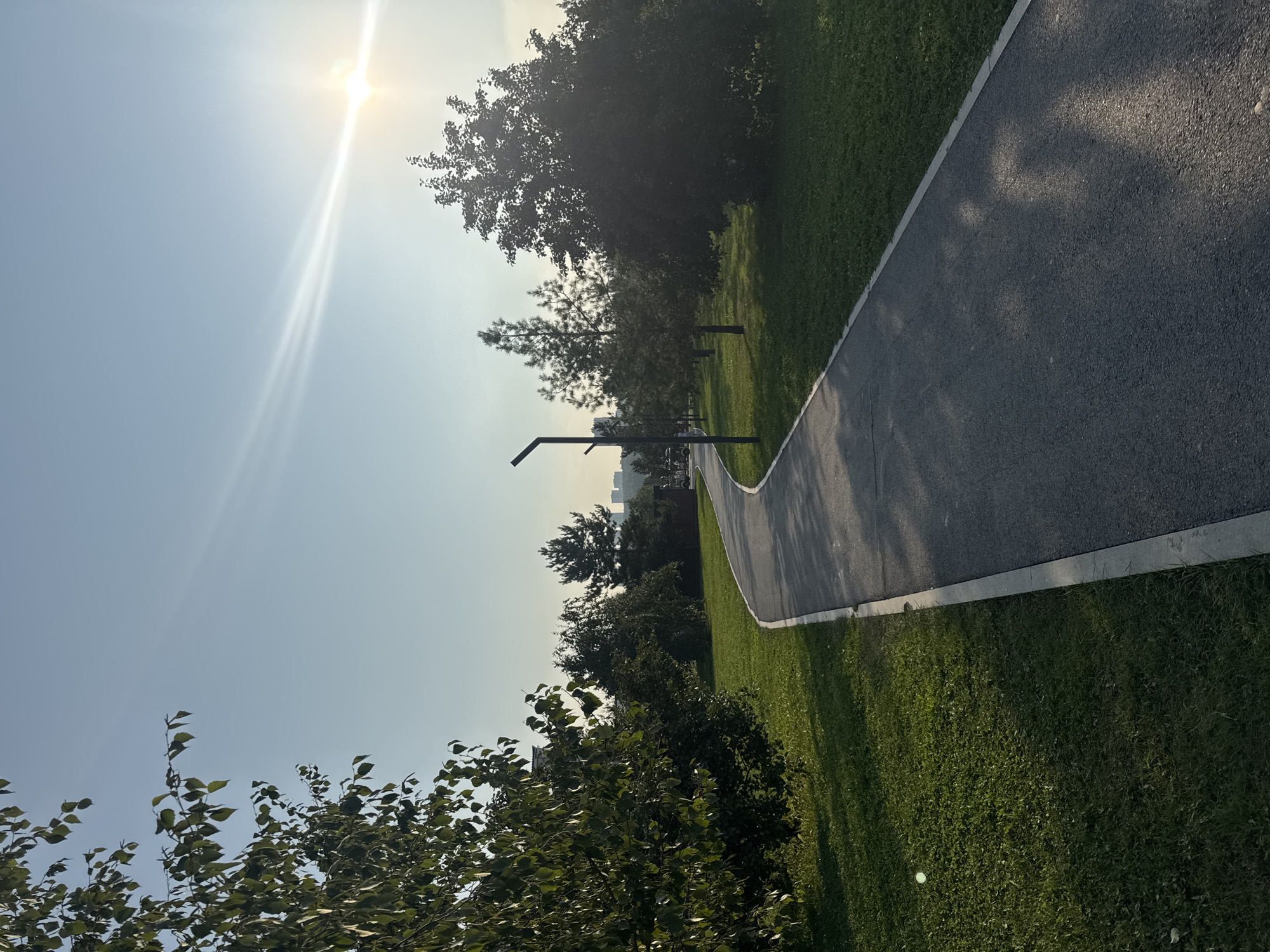 The image shows a paved pathway winding through a grassy park with trees. The sun is visible in the upper right corner, casting a bright glare and creating lens flares. The sky is clear blue with some wispy clouds. In the distance, several buildings can be seen. The pathway is bordered by a concrete edge and is shaded in parts by trees. The grass is green and well-maintained. The scene appears to be set in a public park within the city of Ufa, Russia, likely during daylight hours given the sun's position.
