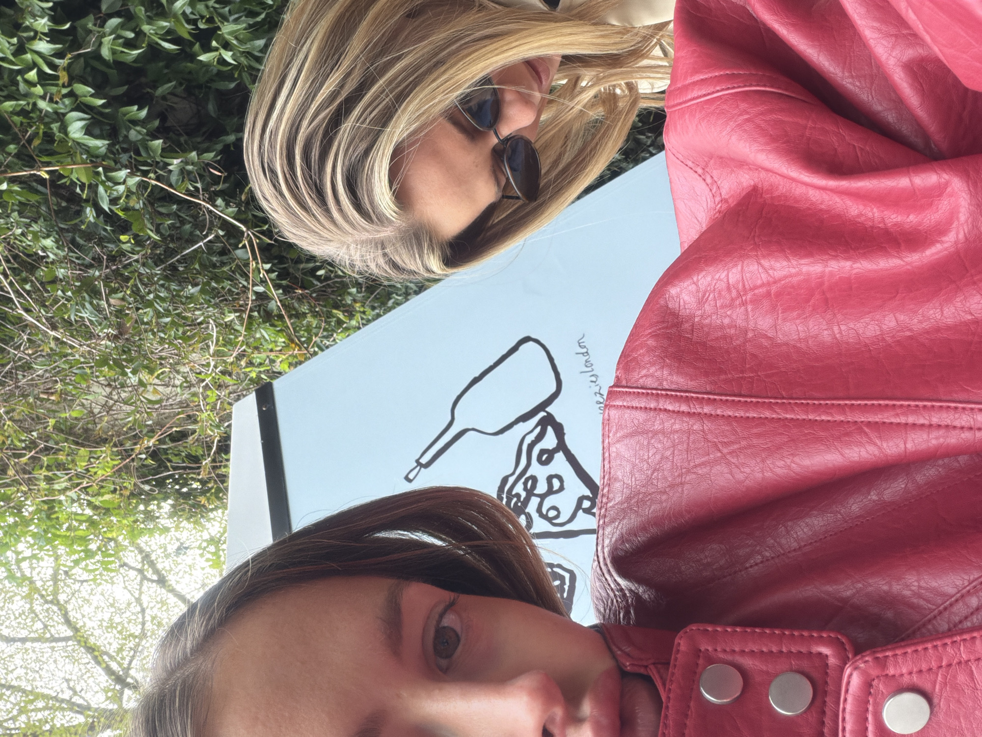 A low-angle selfie, featuring two women standing near a sign, possibly in London, United Kingdom, based on the text visible on the sign. One woman, likely the one holding the camera, is positioned in the foreground. The other woman, with blonde hair and sunglasses, is positioned slightly behind and above her.
The sign between them displays a drawing of what appears to be a bottle and a slice of pizza. The text, partly obscured, includes the word "London."
One woman is wearing a red leather jacket. The background shows a mix of greenery, including trees and bushes.