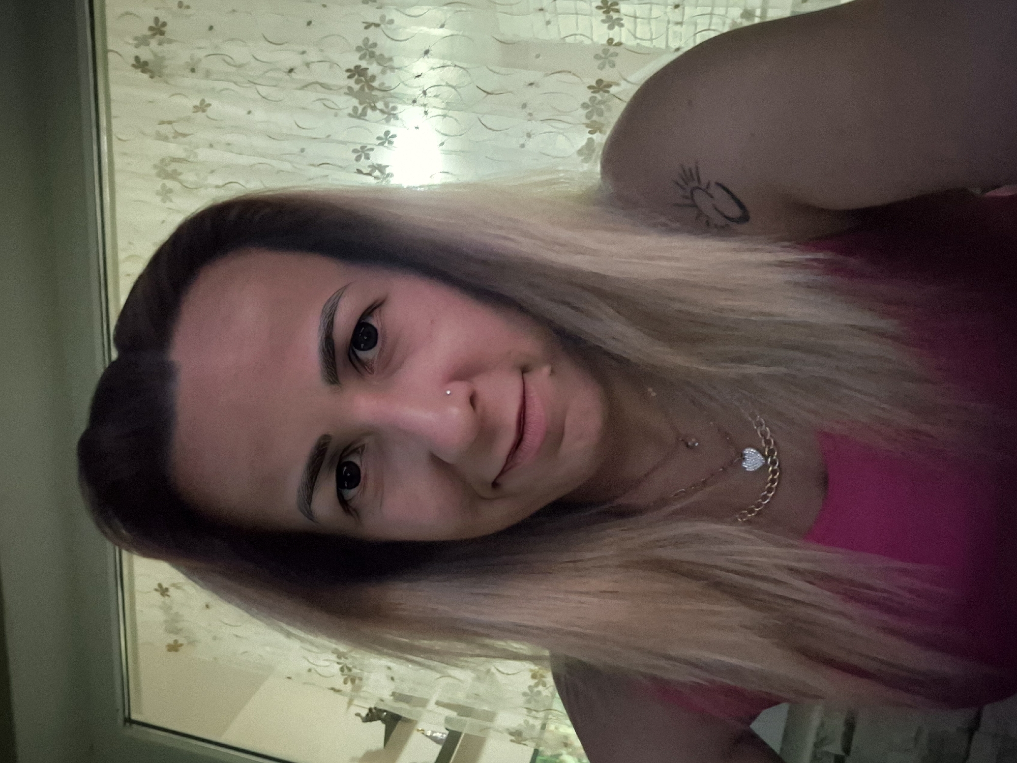 A woman is shown in a close-up selfie. She has blonde hair with darker roots, pulled back slightly from her face. She is wearing a bright pink top and several necklaces, including one with a heart-shaped pendant. There is a small nose ring and a tattoo on her right shoulder. The background appears to be indoors, with a light-colored curtain with a floral pattern behind her. The lighting is somewhat dim, casting shadows on her face. The scene is set in a domestic environment.