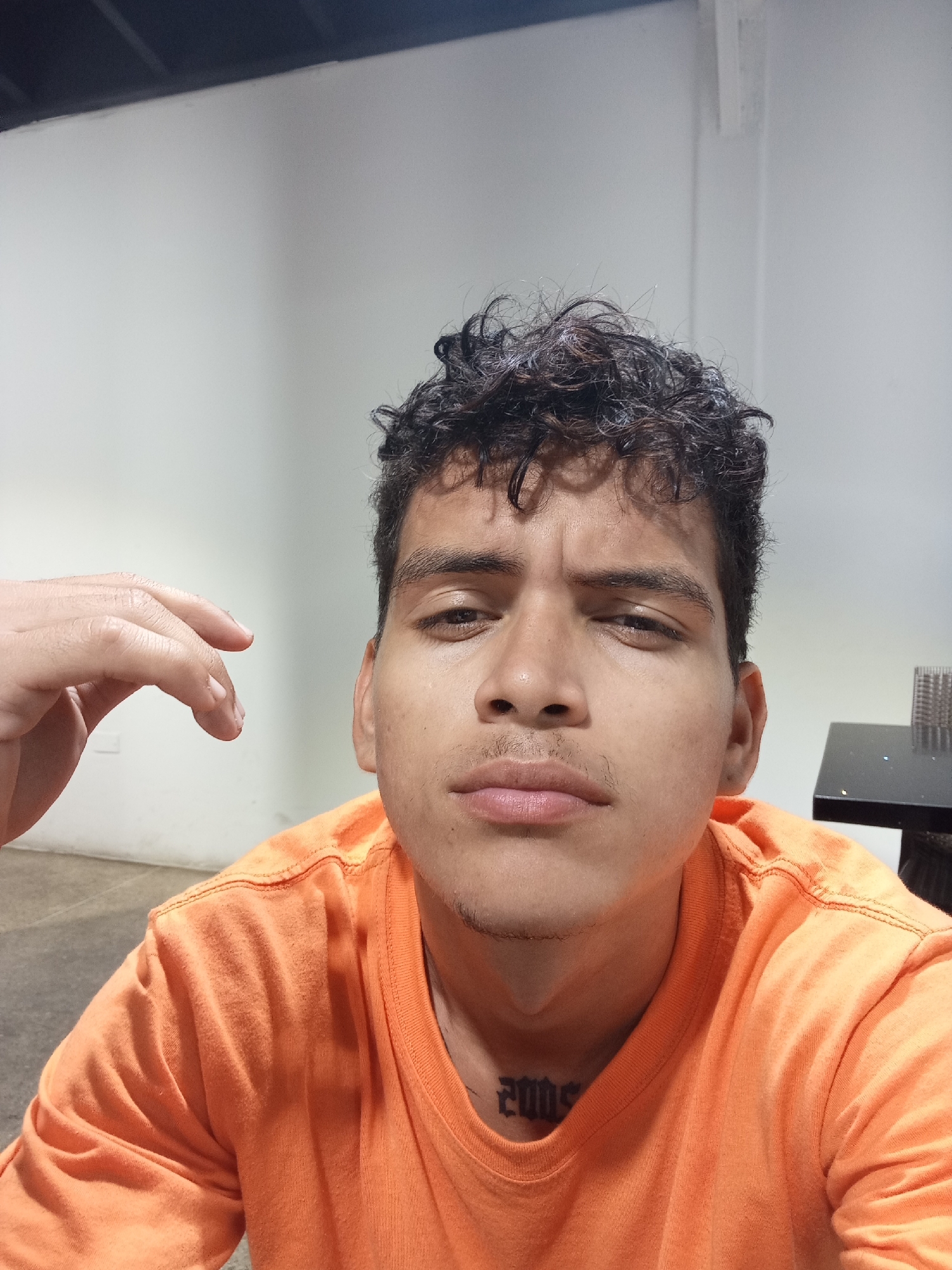 A close-up, selfie-style photograph features a young adult male. He has dark, curly hair, brown eyes, and light brown skin. He is wearing an orange t-shirt. A tattoo, reading "2005," is visible on his neck. Part of his left hand is visible in the left frame.
The background is a plain, white wall, with a dark ceiling fixture visible in the upper left. A dark table with a decorative object is visible in the background to the right.
No explicit location details relating to Juan Griego, Venezuela, are discernible.