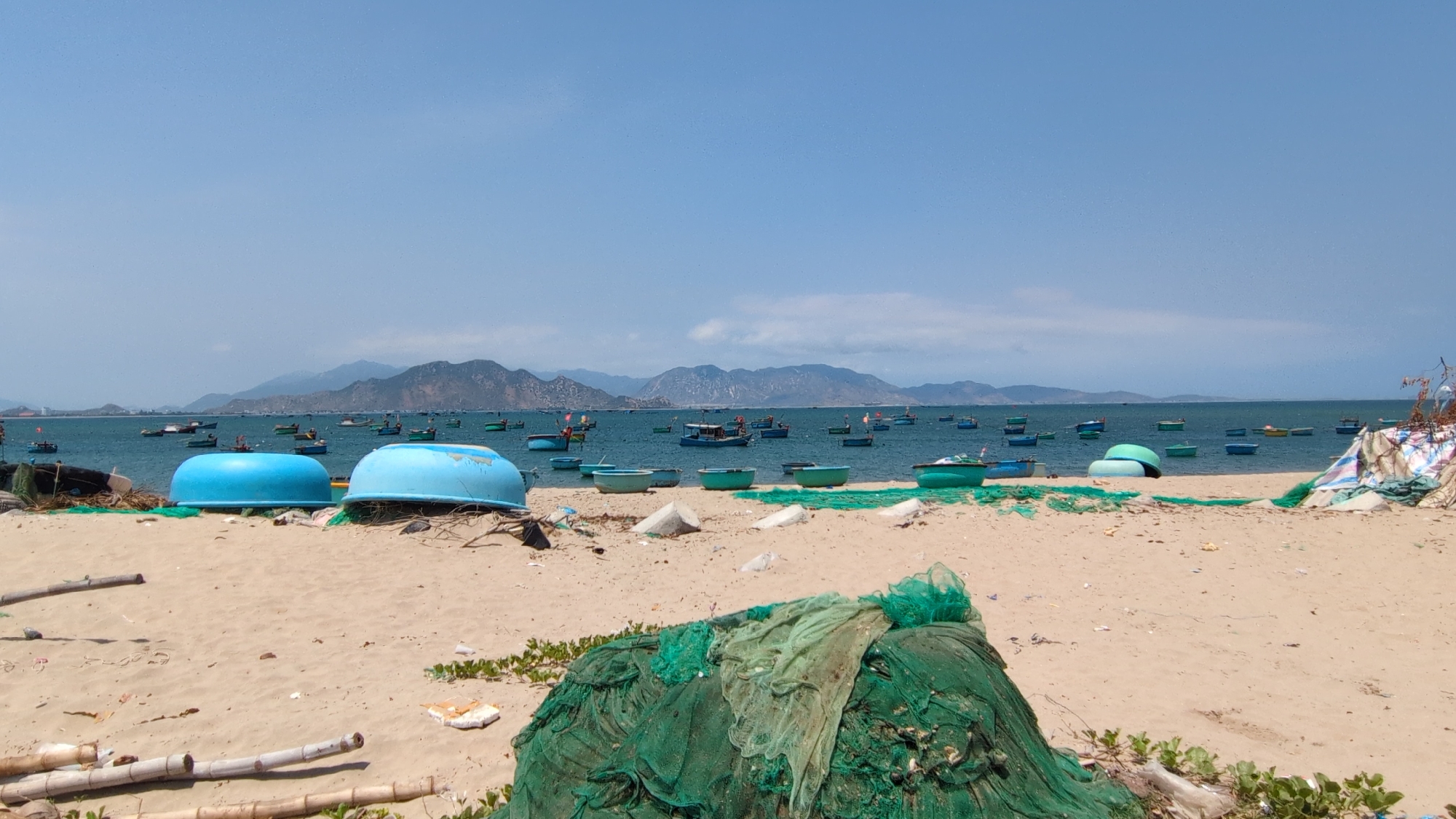 This photograph shows a beach near the city of Phan Rang–Tháp Chàm, Vietnam. The foreground of the scene is dominated by a sandy beach littered with debris, including green fishing nets, bamboo poles, and other miscellaneous items. In the mid-ground, overturned turquoise boats rest on the sand. Beyond the beach is the ocean, filled with numerous small fishing boats. The horizon is marked by a range of mountains under a clear, blue sky.
