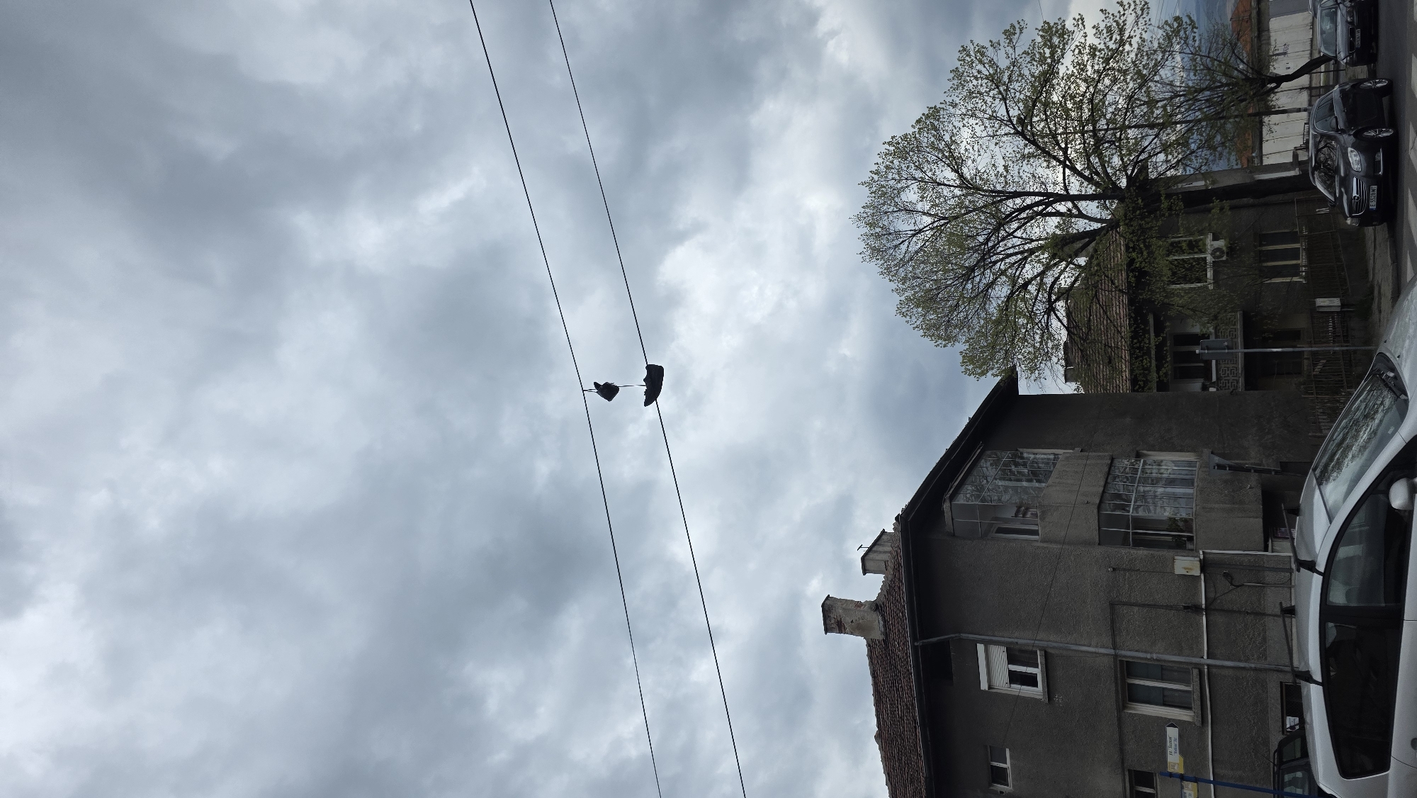 The image shows a pair of shoes hanging from a power line in Blagoevgrad, Bulgaria. The sky is overcast with grey clouds. A building is visible to the right, with a tree partially obscuring it. A car is parked in front of the building.
