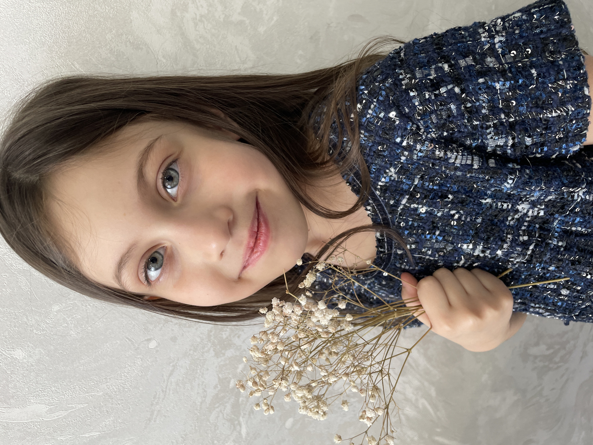 The image presents a head-and-shoulders portrait of a young female subject, estimated to be between 6 and 10 years old, positioned centrally against a light gray, textured wall. The subject has long, straight dark brown hair, fair skin, and light blue-grey eyes, which are directed toward the camera. Her lips are closed in a gentle smile. She is wearing a short-sleeved, round-neck garment, likely a dress or top, made of a dark blue textured fabric with interwoven lighter blue, white, black, and silver threads, along with small, reflective sequins. Her left hand, visible on the lower right side of the frame, holds a small bouquet of dried white or off-white Gypsophila (baby's breath) flowers with thin, light brown stems. The background is a nondescript interior wall, offering no specific visual identifiers for a location within Izhevsk, Russia, as specified.