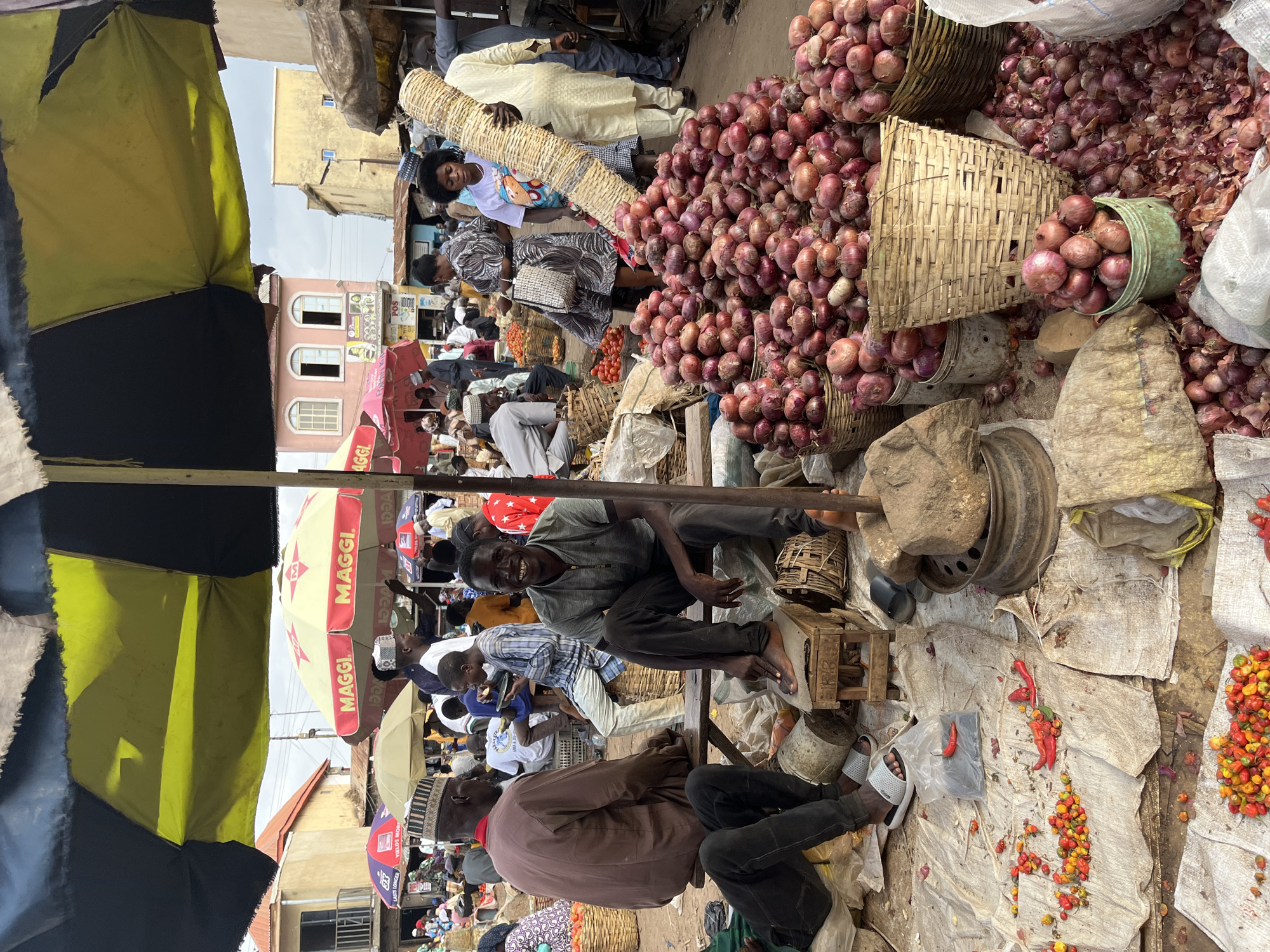 The image depicts a bustling outdoor market scene in Rafin Par, Nigeria. The foreground is dominated by large piles of red onions, displayed in woven baskets and buckets, with some onions scattered on the ground. To the left, a collection of small, colorful chili peppers are spread across a white cloth. Several individuals are present, some sitting on wooden crates or benches, others standing and carrying goods.
A man in a grey t-shirt and dark pants is seated prominently in the center, smiling at the camera. To his left, another man in a brown tunic and patterned cap is also seated. In the background, a woman carries a large, cylindrical woven basket on her shoulder. Other marketgoers are visible amidst stalls and produce, under the shade of large umbrellas. The environment suggests a typical marketplace with a variety of goods for sale, likely food items. The overall impression is one of vibrant commerce and community activity.