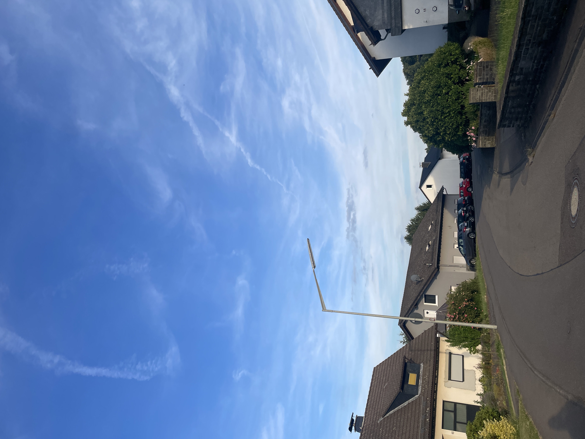 The image depicts a quiet residential street in Wiehl, Germany, under a bright blue sky with wispy white clouds and contrails. A paved road curves through the scene, lined with several houses of varying architectural styles. On the left, a light-colored house with a dark-tiled roof and a dormer window is visible. To its right, a grey house with a satellite dish is situated, with a small rose bush in front. Further down the street, more houses and parked cars, including a dark grey hatchback and a red car, are present. A tall, modern street lamp with a single light fixture stands prominently in the middle ground, casting no discernible shadow. A dense green tree dominates the right side of the frame, with a low brick wall running along the edge of the street in front of it. The overall impression is one of a peaceful suburban environment during daylight hours.