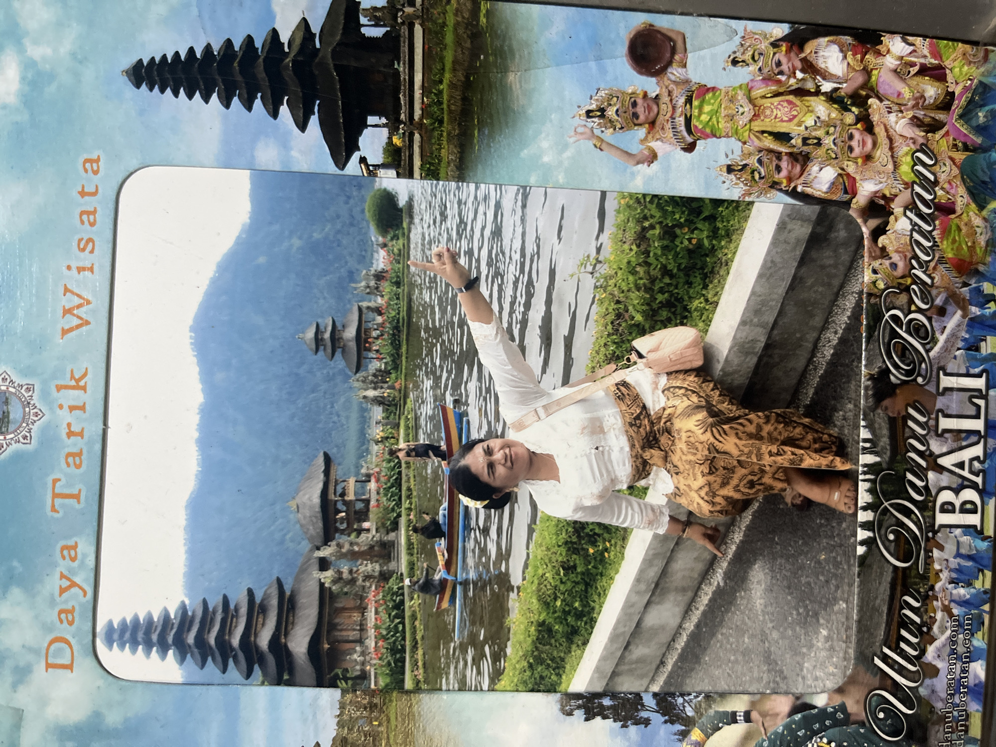 This detailed image depicts a composite promotional postcard or souvenir frame showcasing the Pura Ulun Danu Beratan temple in Mendoyo, Indonesia. The central and dominant feature is a photograph framed with rounded corners. In this central photo, a woman with dark hair styled with a yellow flower is seated on a grey concrete ledge at the edge of a body of water, possibly Lake Bratan. She wears a white, long-sleeved blouse with lace and a patterned brown and gold traditional sarong, along with a light pink cross-body bag. Her right arm is raised, with her index finger pointing upwards, and she is smiling towards the viewer.
Behind the woman, the calm water reflects light. A small, multi-colored boat (red, blue, yellow) is on the water, containing two men, one of whom appears to be paddling. On the far bank of the water, several traditional Balinese temple structures are visible, including a prominent multi-tiered meru (pagoda-like tower) and smaller thatched pavilions, surrounded by lush green foliage and patches of red flowers. In the distant background, hazy blue mountains rise under a light blue sky.
Surrounding this central framed photograph are additional decorative elements. Across the top, the orange text "Daya Tarik Wisata" is visible against a light blue, cloudy sky background. The lower section features large, ornate white script spelling "Ulun Danu Beratan BALI," with a smaller website address "danuberatan.com" below it. On the right side of the frame, a vertical strip displays another view of a multi-tiered temple structure by water. Below this, a group of Balinese dancers in elaborate gold, green, and pink traditional costumes with intricate headdresses are shown. One dancer stands elevated above the others, holding a small, round, brown object, possibly a bowl or drum. On the far left, a narrow vertical strip shows another partial view of temple architecture and water. The overall impression is a blend of a personal travel photo with iconic cultural and scenic elements of the location.