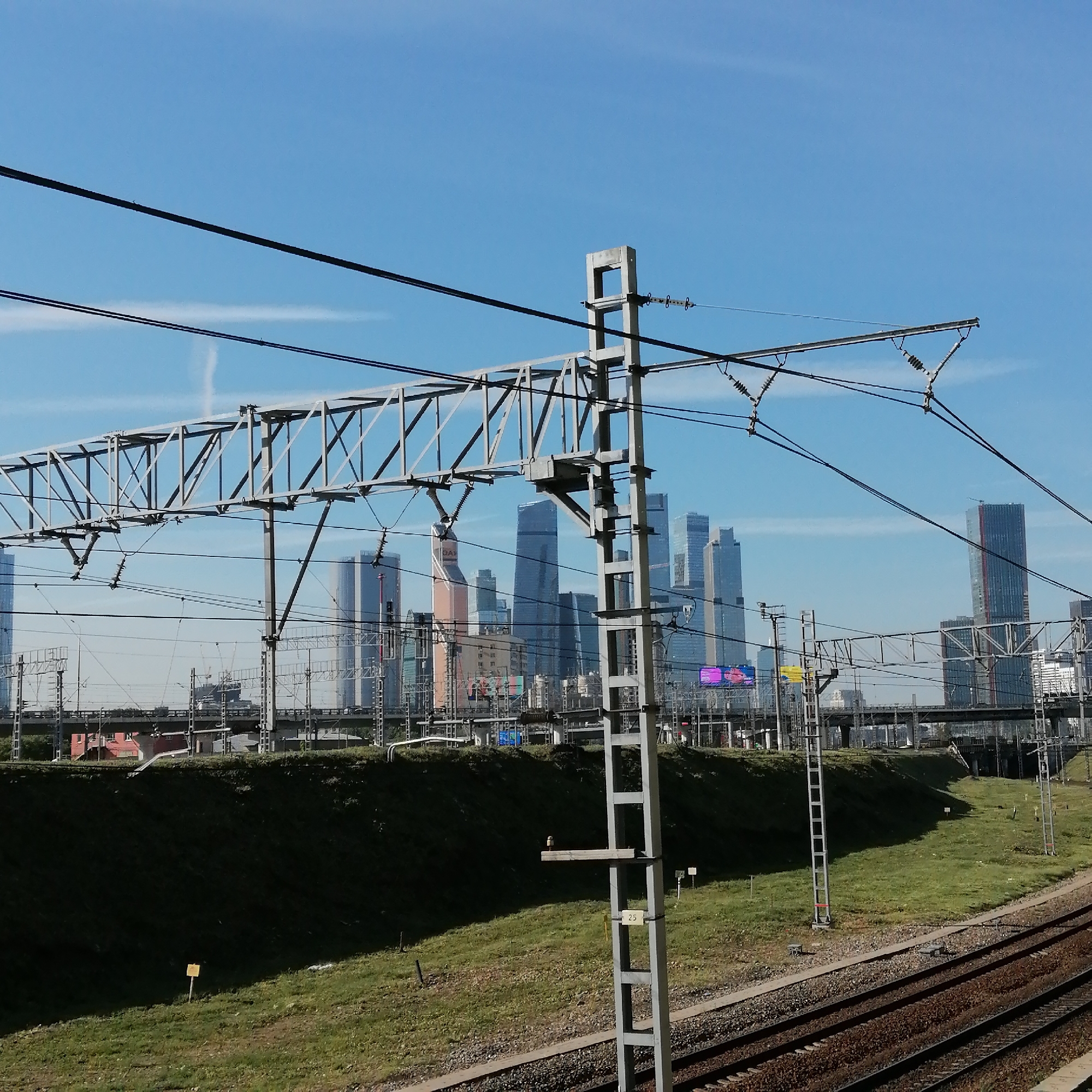 The image shows a view of the Moscow International Business Center skyline from a ground-level perspective along a railway track. The sky is clear and blue. In the foreground, there are railway tracks running along the bottom right corner of the frame, alongside a grassy embankment. A gray, lattice-like metal structure, part of the railway's infrastructure, is positioned to the left of the foreground. Behind the embankment, the cityscape of Moscow is visible, featuring a cluster of modern skyscrapers of varying heights and architectural designs. The image is taken in daylight and the overall composition suggests an urban landscape viewed from a railway line on the outskirts of the city center.