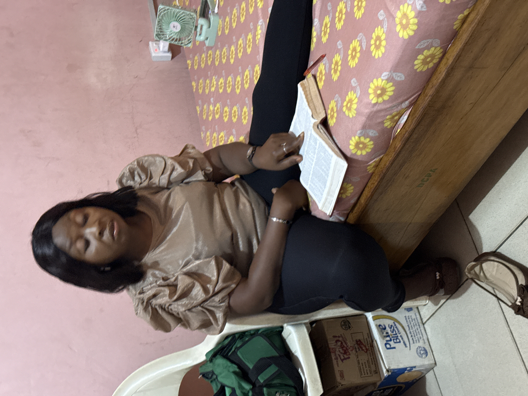A woman of African descent is lying on a bed, reading a book with a red pen inside. She is wearing a tan-colored blouse and black leggings. Her left arm rests on the book, while her right hand is placed on the open pages. The bed has a pink patterned bedspread with yellow flowers, and the headboard is made of wood. A small green electric fan is positioned on the bed. She is laying on top of several boxes. A pair of brown flats are on the floor. The wall is pink. It is unknown if the location is in Abuja, Nigeria.
