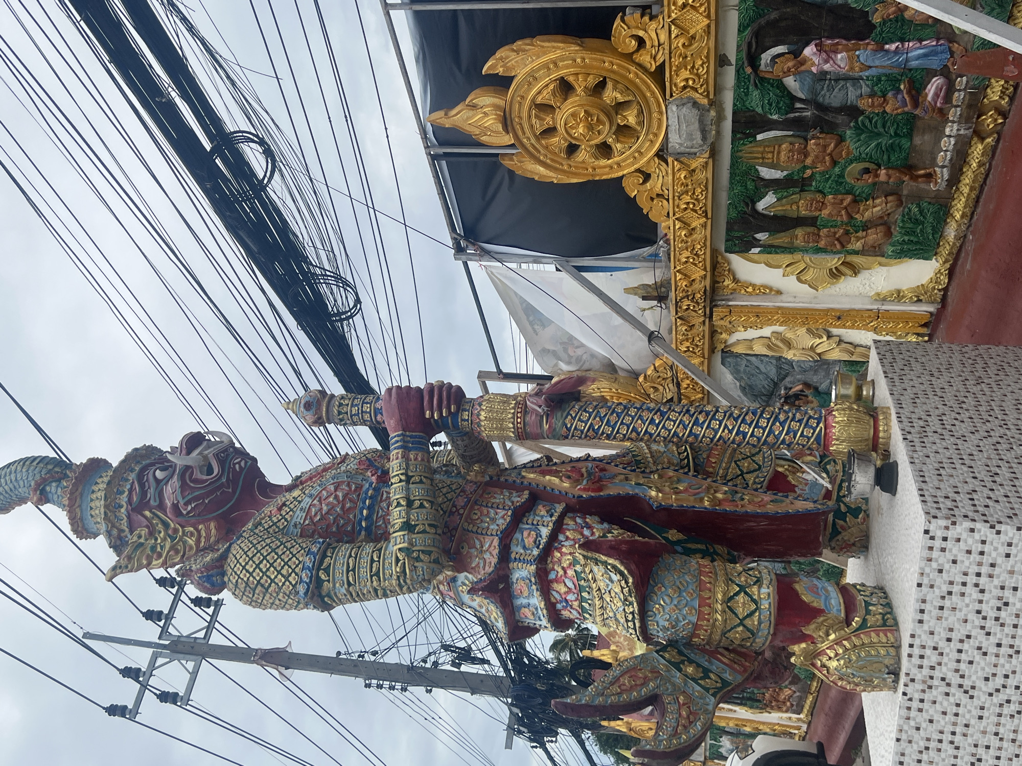 In an outdoor setting under a cloudy sky, a large, intricately decorated statue of a mythological creature stands prominently. The statue is predominantly red with elaborate gold, blue, and green decorative patterns. It depicts a fierce-looking being with a long snout, tusks, and an ornate headdress. The creature holds a staff or club in its hands.
To the right of the main statue, there is a decorative golden wheel with flames, a symbol often associated with Buddhism. Below this, a relief sculpture on a wall depicts several figures in a natural setting with trees. One figure in the foreground wears a pink and blue garment.
The scene is characterized by an abundance of overhead electrical wires, suggesting an urban or semi-urban environment. A power pole is visible to the left of the main statue. The base of the main statue is adorned with small, light-colored mosaic tiles. The overall impression is one of religious or cultural ornamentation.