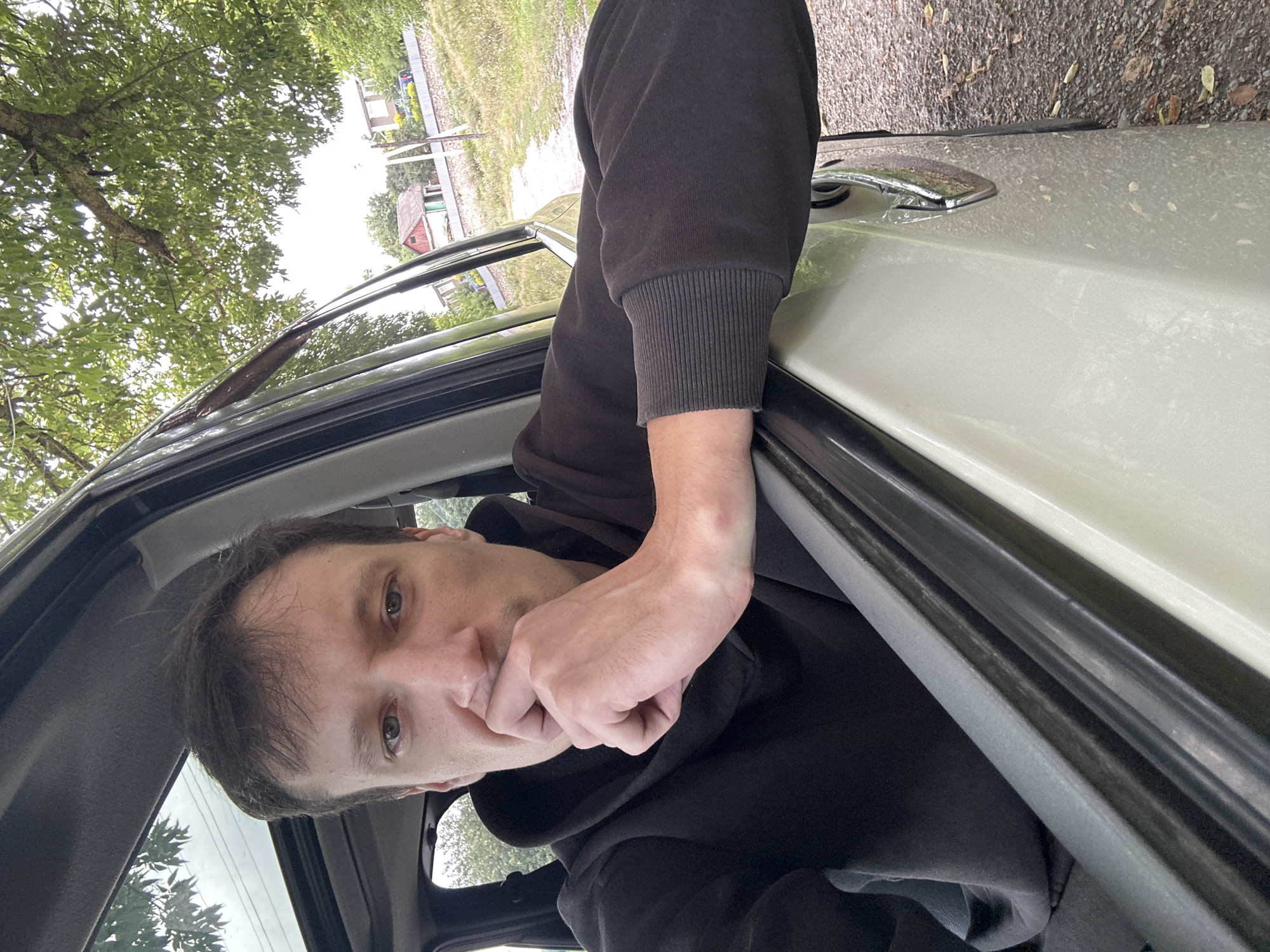 A medium shot shows a man inside a car, with his left arm resting on the open window frame and his hand covering his mouth, looking directly at the camera. He is wearing a black hooded sweatshirt. The car interior is visible with a sunroof open above. Outside the car, a rural landscape is depicted with trees, a dirt road, a stone fence, and houses in the distance. The scene appears to be in Makiv, Ukraine, given the style of houses and the natural surroundings. The lighting suggests it is daytime.