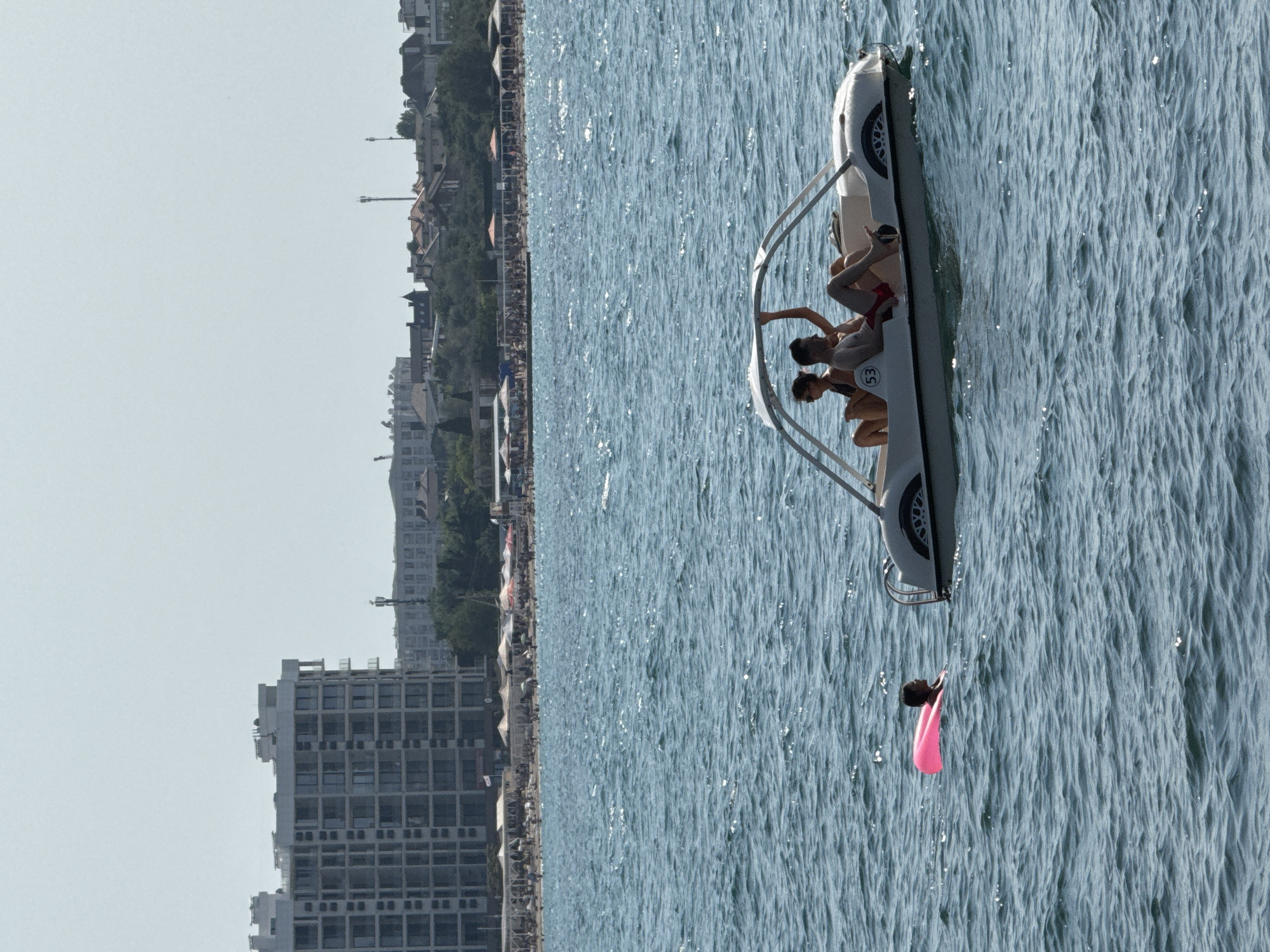In the foreground of the image, the choppy blue water of the sea is visible. In the mid-ground, a white pedal boat with a canopy is in motion. Inside the boat, two individuals, a male and a female, are seated. The male is wearing red shorts, and the female is wearing a dark-colored swimsuit. To the left of the pedal boat, a person wearing a dark-colored swimsuit is floating on a bright pink inflatable tube.
In the background, a crowded beach is visible with numerous people and beach umbrellas. Behind the beach, there are several multi-story buildings, including a large, modern building with many windows. The cityscape extends further into the distance, with more buildings and trees dotting the horizon under a clear, hazy sky. The scene is likely from a coastal area in Sevastopol, Ukraine, given the architecture and the presence of beachgoers enjoying water activities.
