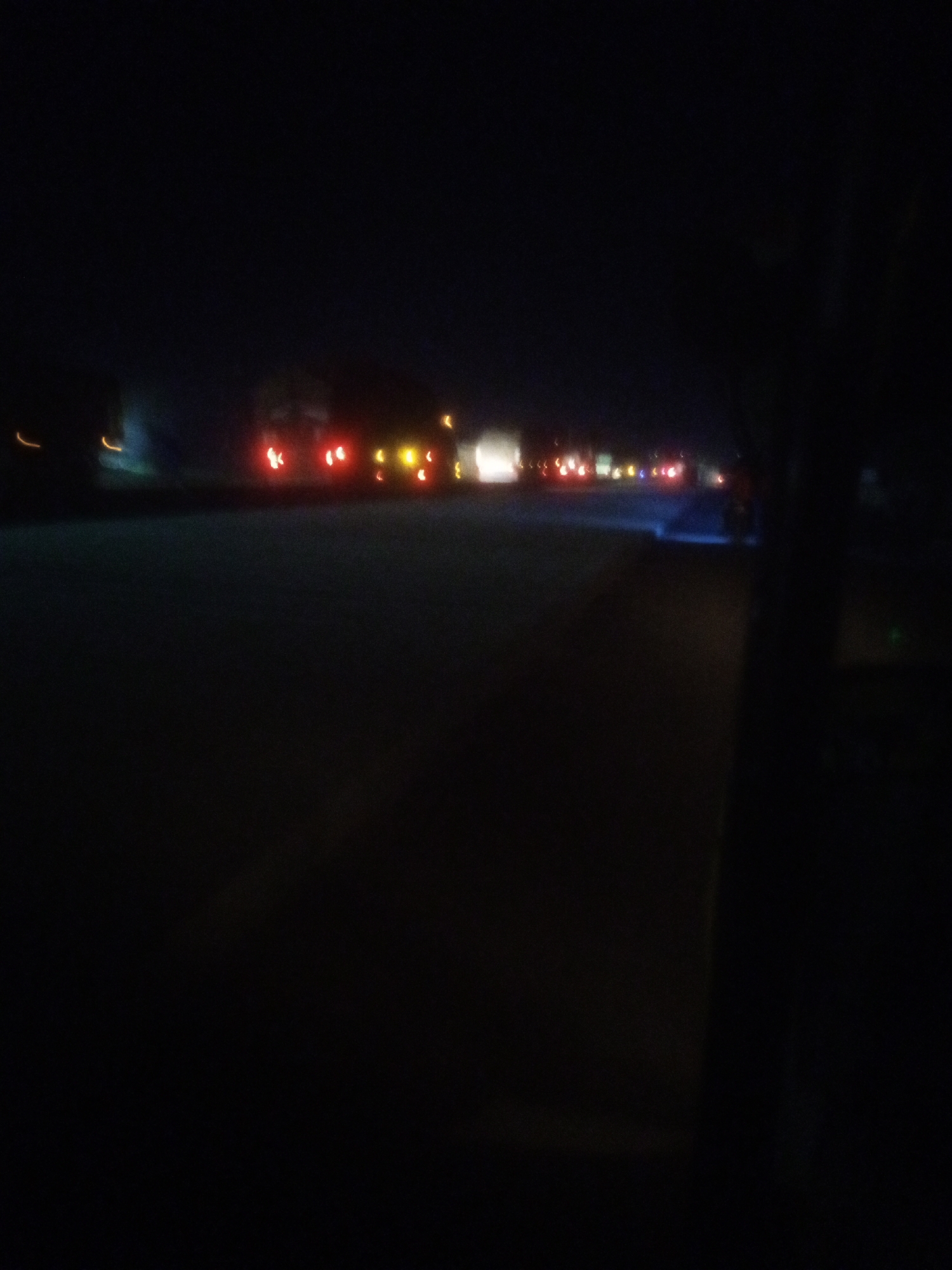 The image depicts a night scene on a road, possibly in or near Nassarawa, Nigeria, characterized by motion blur and low light conditions. Several vehicles are visible, indicated by their illuminated taillights, appearing as streaks of red and yellow. These lights suggest vehicles are in motion, likely on a highway or main road. The environment is dark, with only the vehicle lights and faint ambient light providing illumination. The foreground is predominantly dark, with the road surface barely discernible. The overall impression is that of a moving vehicle capturing the scene at night.