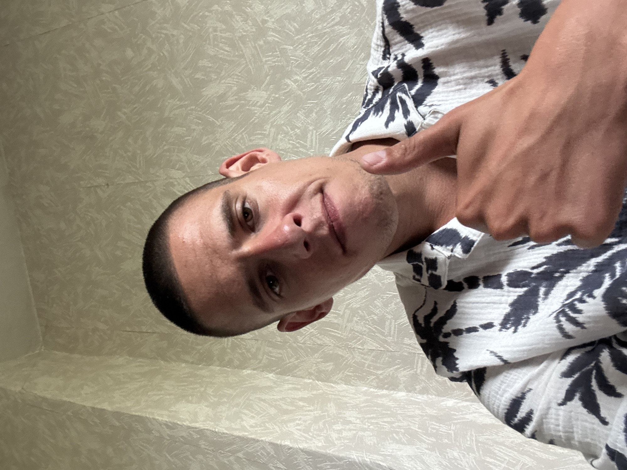 A man is looking down at the camera and pointing at his chin with his right index finger. He has short dark hair, light skin, and stubble. He is wearing a short-sleeved white shirt with a dark pattern of leaves. The background is a light-colored wall with a textured pattern. The image does not contain sufficient information to identify a specific location in Sofia, Bulgaria.