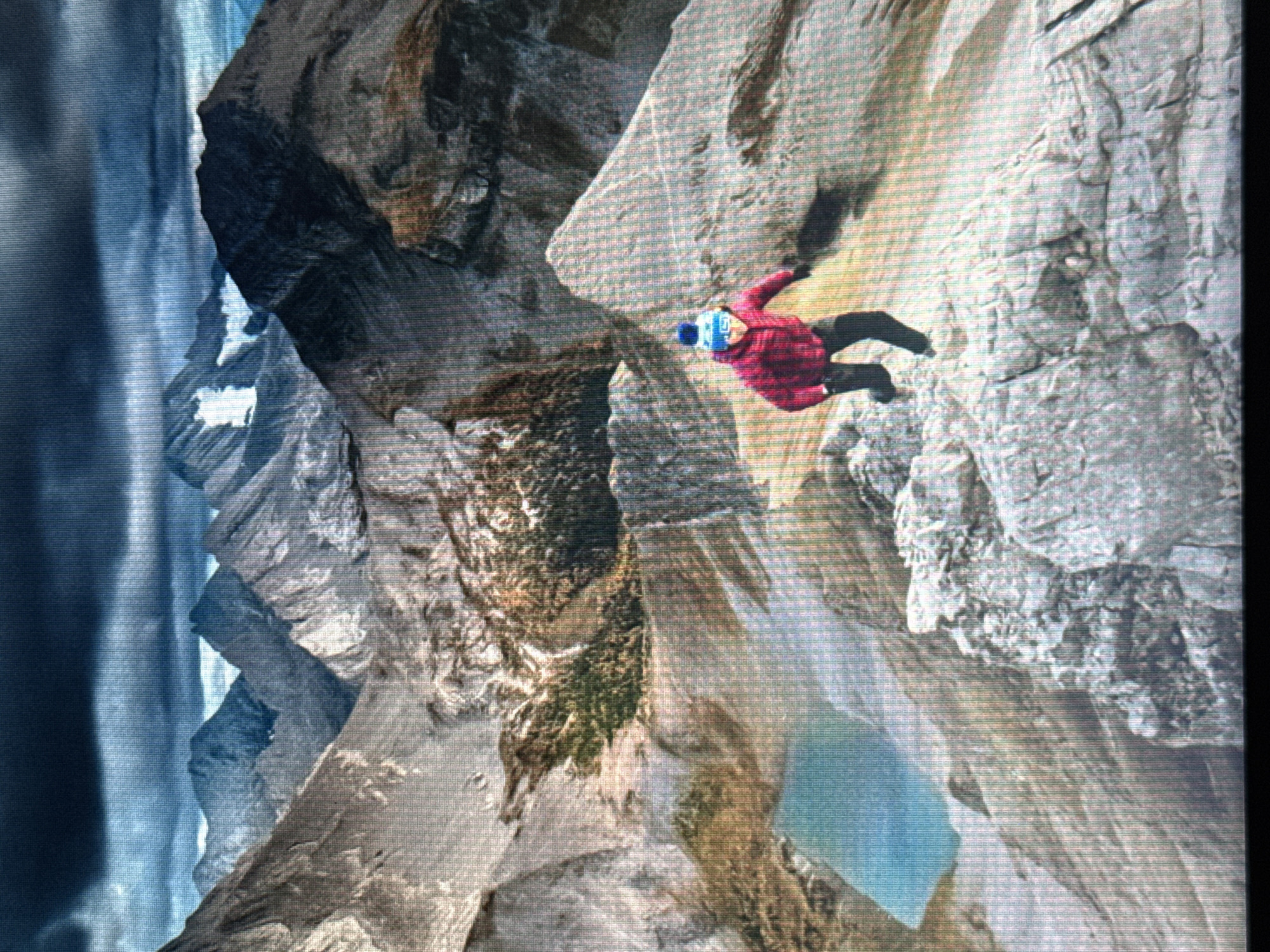 This is a photograph of a person wearing a red jacket and a blue and white pom-pom hat, running down a rocky mountain slope. The person is wearing dark pants. The background shows a dramatic mountain landscape with dark, cloudy skies. A small, bright blue lake is visible in the mid-ground. The image appears to be taken from a high vantage point, looking down on the scene. The photograph is not of the city of Norilsk, Russia; it depicts a mountainous natural environment.