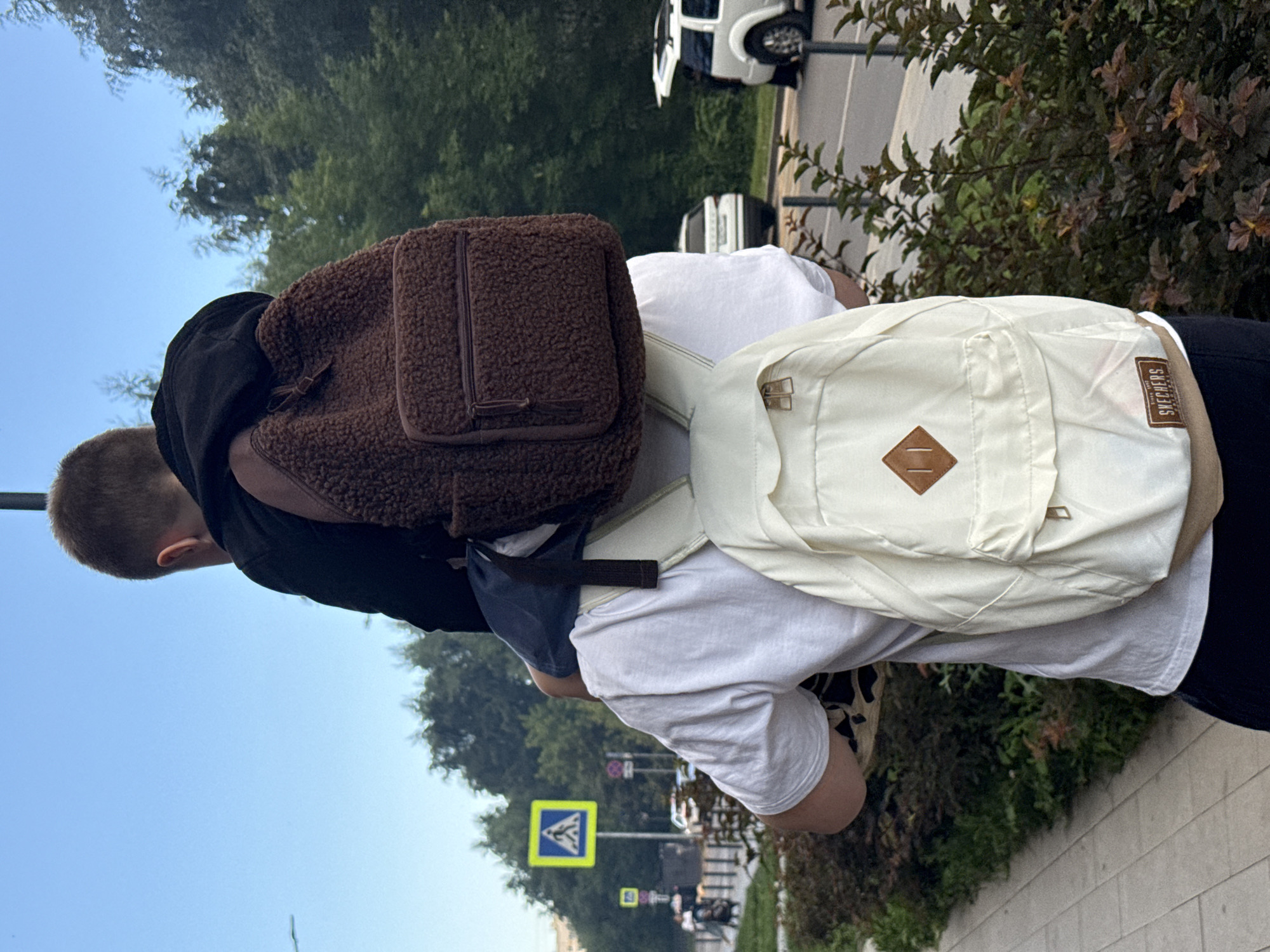 In Lyubertsy, Russia, a person is carrying another person on their shoulders, both wearing backpacks. The person carrying has a white t-shirt and dark pants, with a light-colored backpack on their back. The person on their shoulders wears a black hoodie and a brown, fuzzy backpack. They are walking along a sidewalk next to a road with trees and buildings in the background. A pedestrian crossing sign is visible to the left.