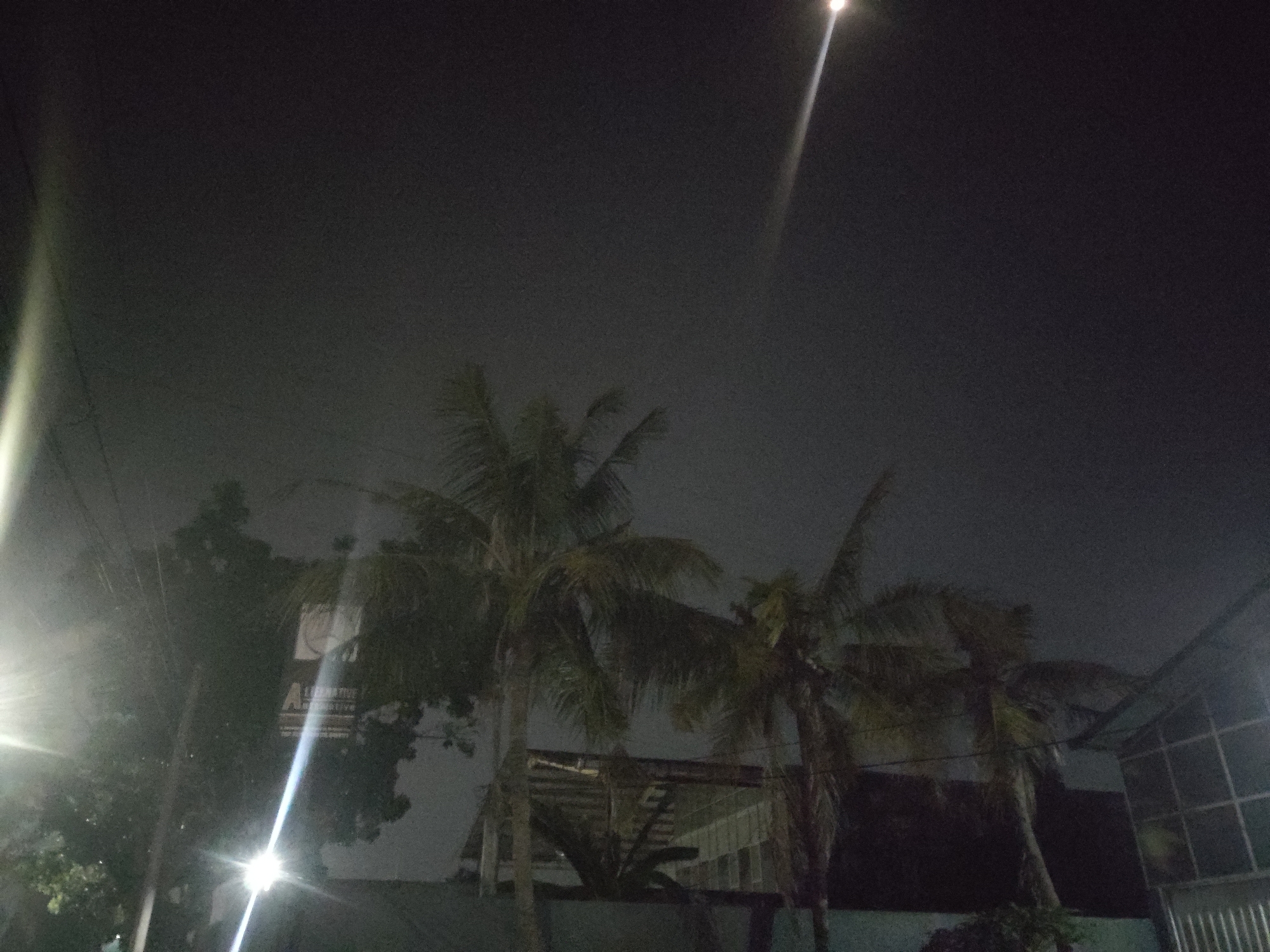 In Trihanggo, Indonesia, at night, a street scene is captured under a dark, hazy sky. A bright light beam emanates from the upper left, possibly from a camera flash or a powerful light source, streaking across the frame. Another bright light flares from the lower left, illuminating a streetlamp or similar fixture. Several palm trees are visible, their fronds dark against the night sky, some appearing to be swaying. In the mid-ground, a building with a tiled roof is partially obscured by the trees. A sign with the letter "A" is visible on the left side, with some unreadable text below it. The overall impression is a dimly lit, somewhat humid night in a tropical urban or suburban setting.