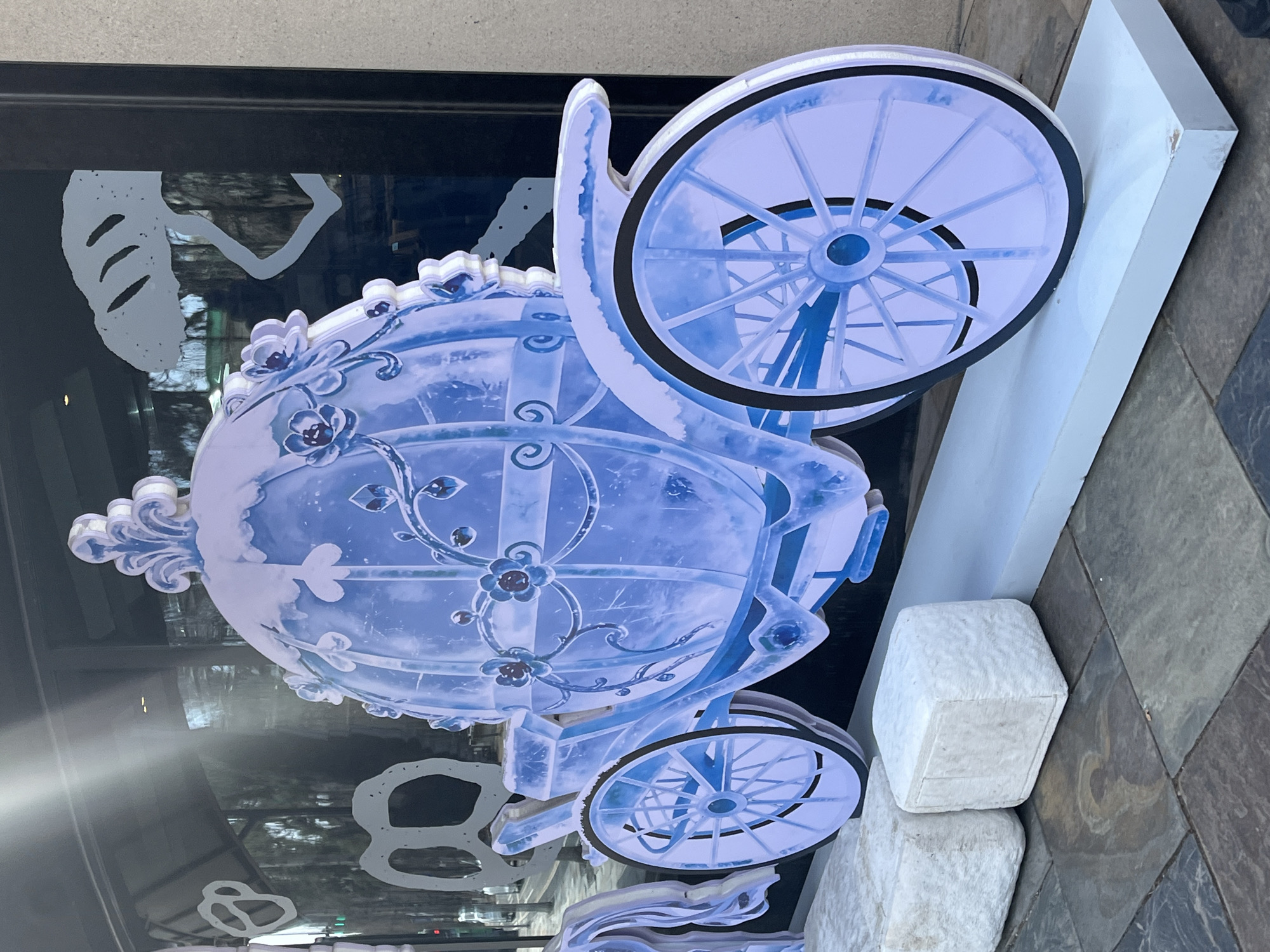 A fairytale-themed display is situated outside a building with a glass facade. The display features a large, ornate, light-blue carriage with detailed floral designs and large wheels. The carriage is mounted on a white base and supported by white blocks underneath. The glass facade behind the display reflects the surrounding environment. The ground around the display is paved with square tiles. The location is Changchun City, China.