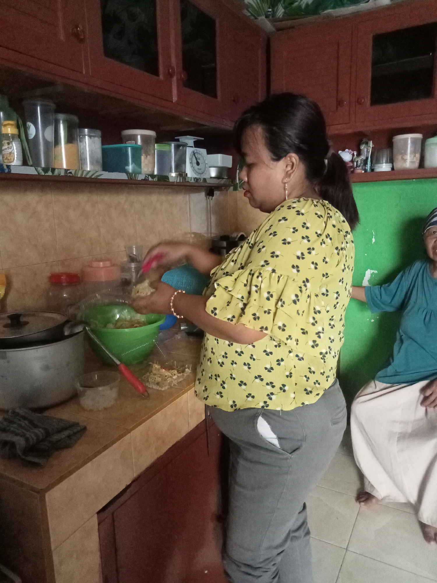 In a kitchen in Malang, Indonesia, a woman with dark hair tied in a ponytail is preparing food. She wears a yellow blouse with a floral pattern and grey pants. Her right hand holds a red utensil, and she is adding ingredients to a green bowl. On the counter, a large pot, a small container of white granules, and other kitchenware are visible. To the right, another woman wearing a teal shirt and a headscarf sits on the floor, her lower body visible. The kitchen features wooden cabinets, tiled walls, and a green painted wall in the background.