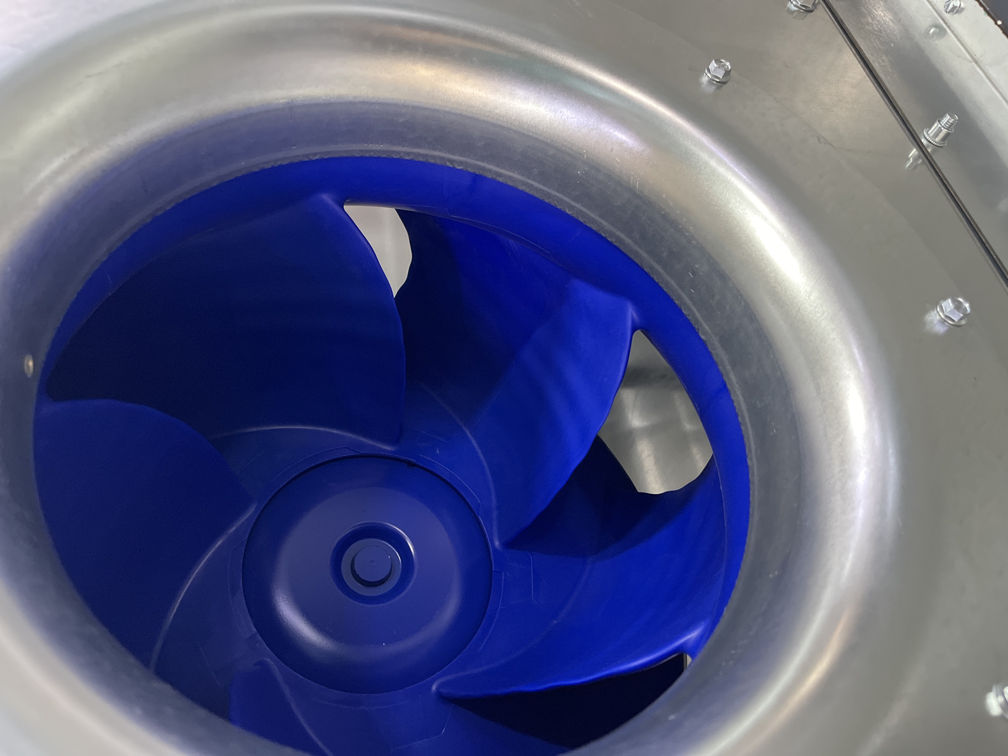The image shows a close-up, angled view of the interior of a centrifugal fan, likely part of an industrial ventilation system. The fan blades are a vibrant blue plastic and are curved, designed to move air. They are housed within a brushed metal casing, which appears to be stainless steel or aluminum. Several bolts are visible securing the metal housing. The lighting creates reflections on the metal surface, highlighting its texture. There are no people or specific landmarks visible that would indicate a location within Kyiv, Ukraine. The scene is sterile and industrial in nature.